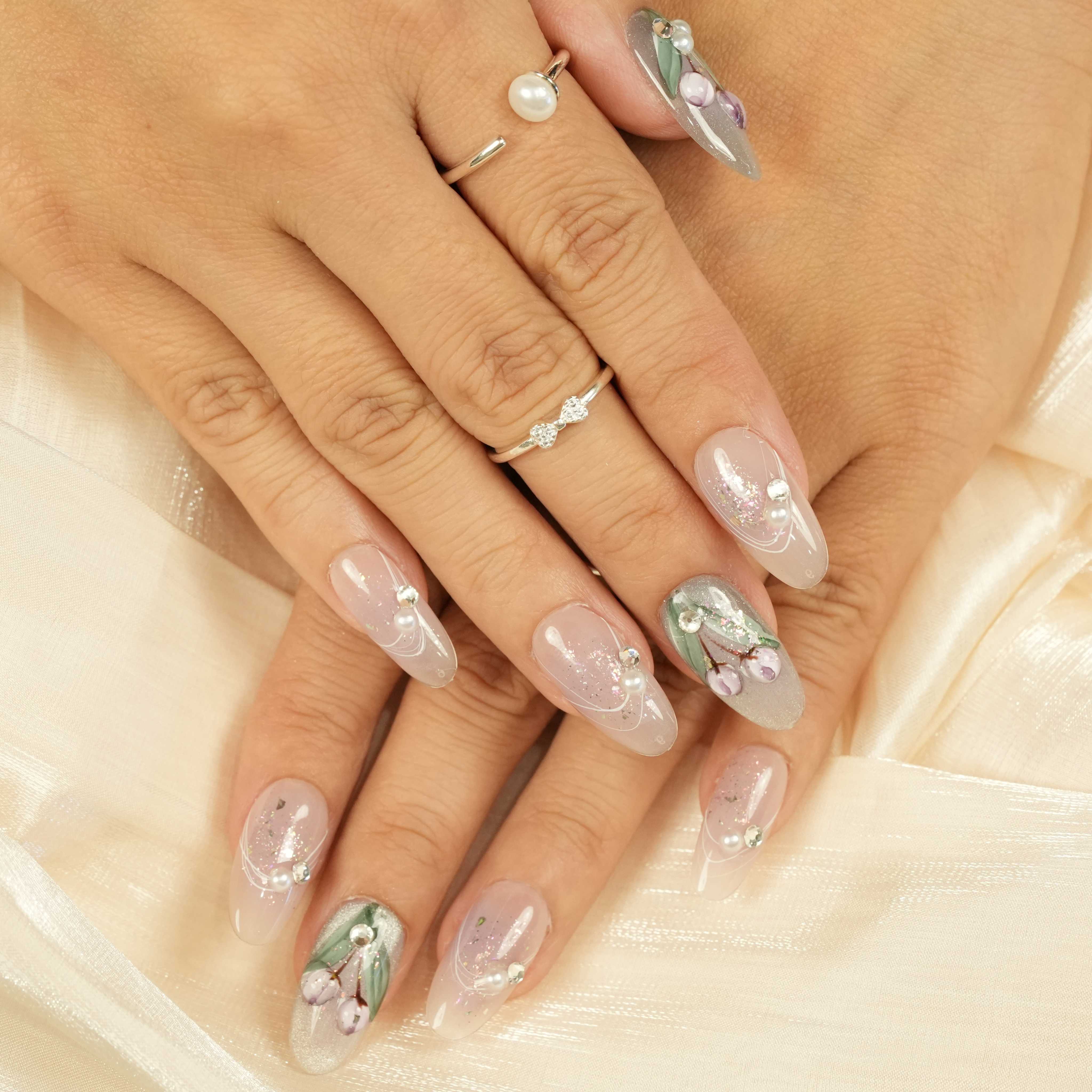 Flat lay of press on nails featuring soft nude and sage green tones with delicate purple floral accents, pearl and rhinestone embellishments