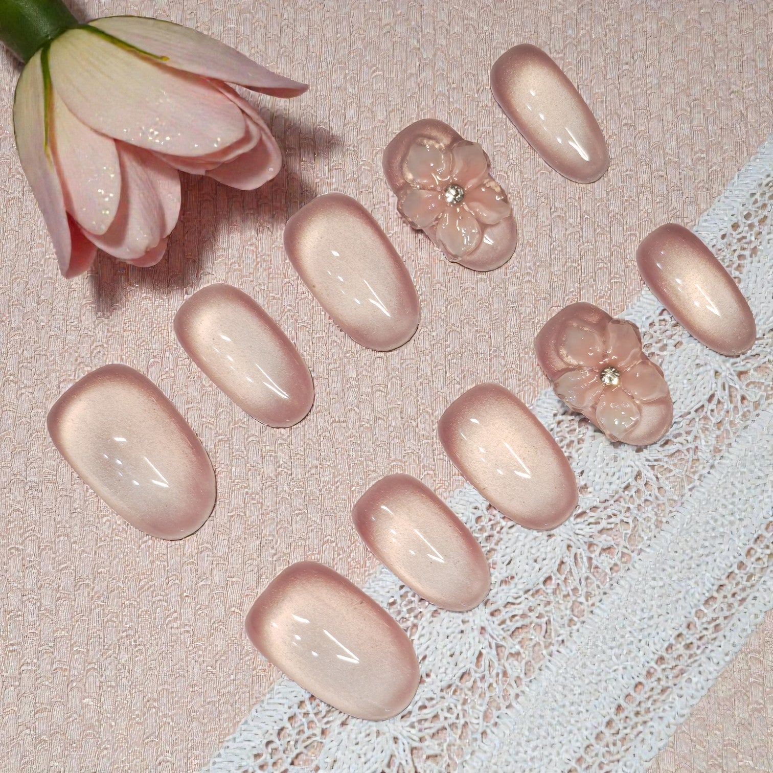 Elegant nude shimmer nail press on set with sculpted 3D flower accents, styled on lace and soft pink fabric for a romantic display.