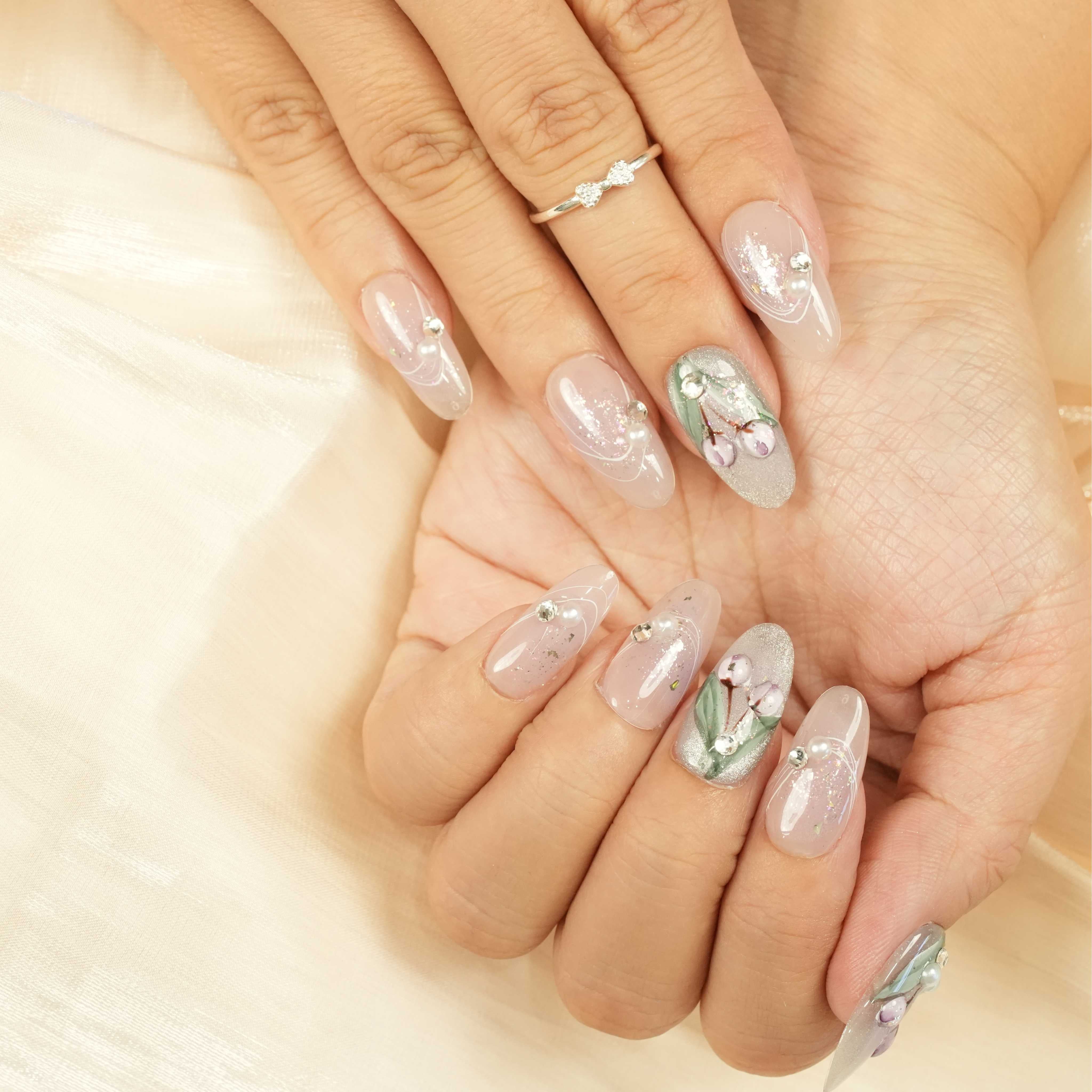 Both hands displaying floral press on nails with nude and green tones, purple blossom art, glitter and pearl details