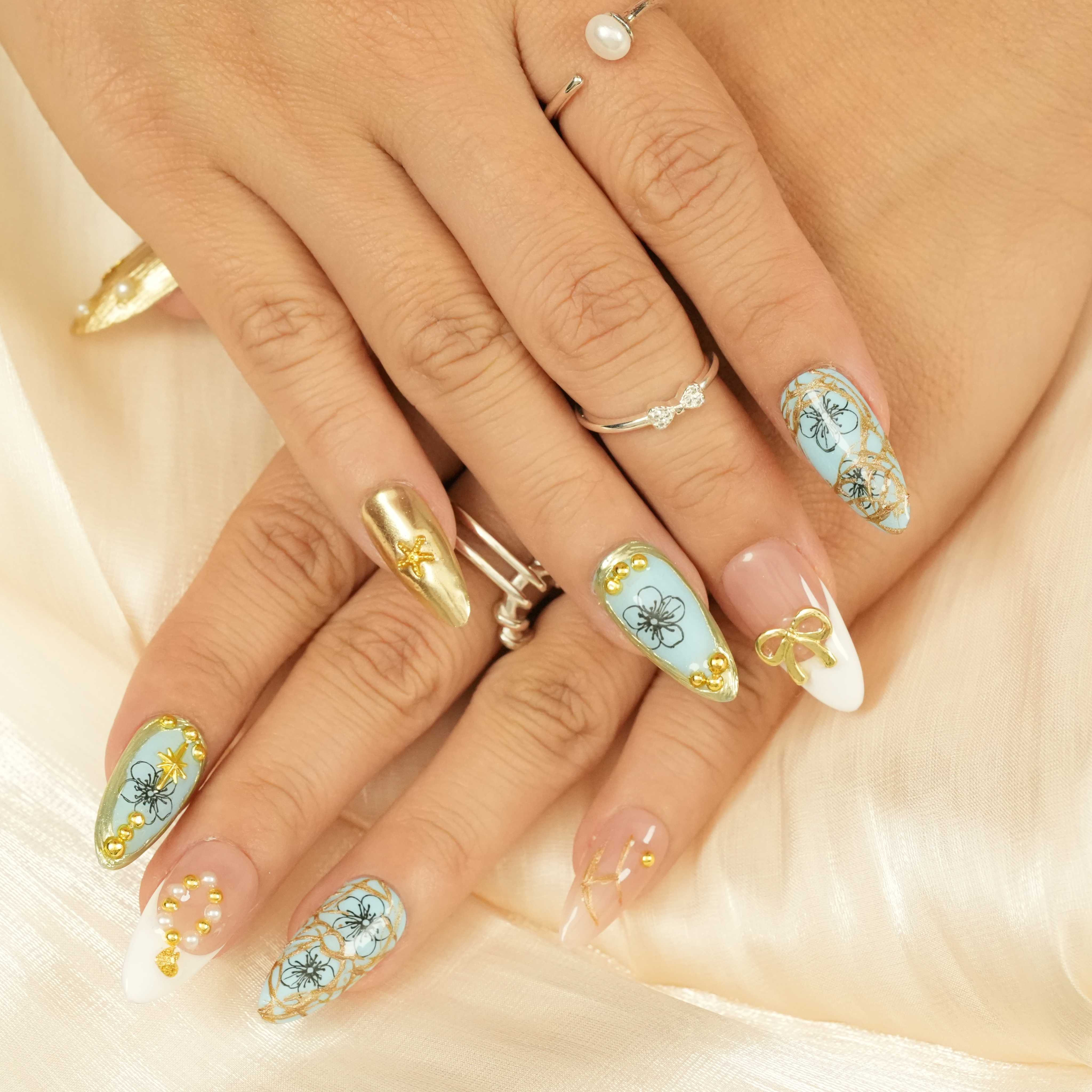 Hand pose highlighting detailed blue and gold floral press on nails with pearls and gold ribbon