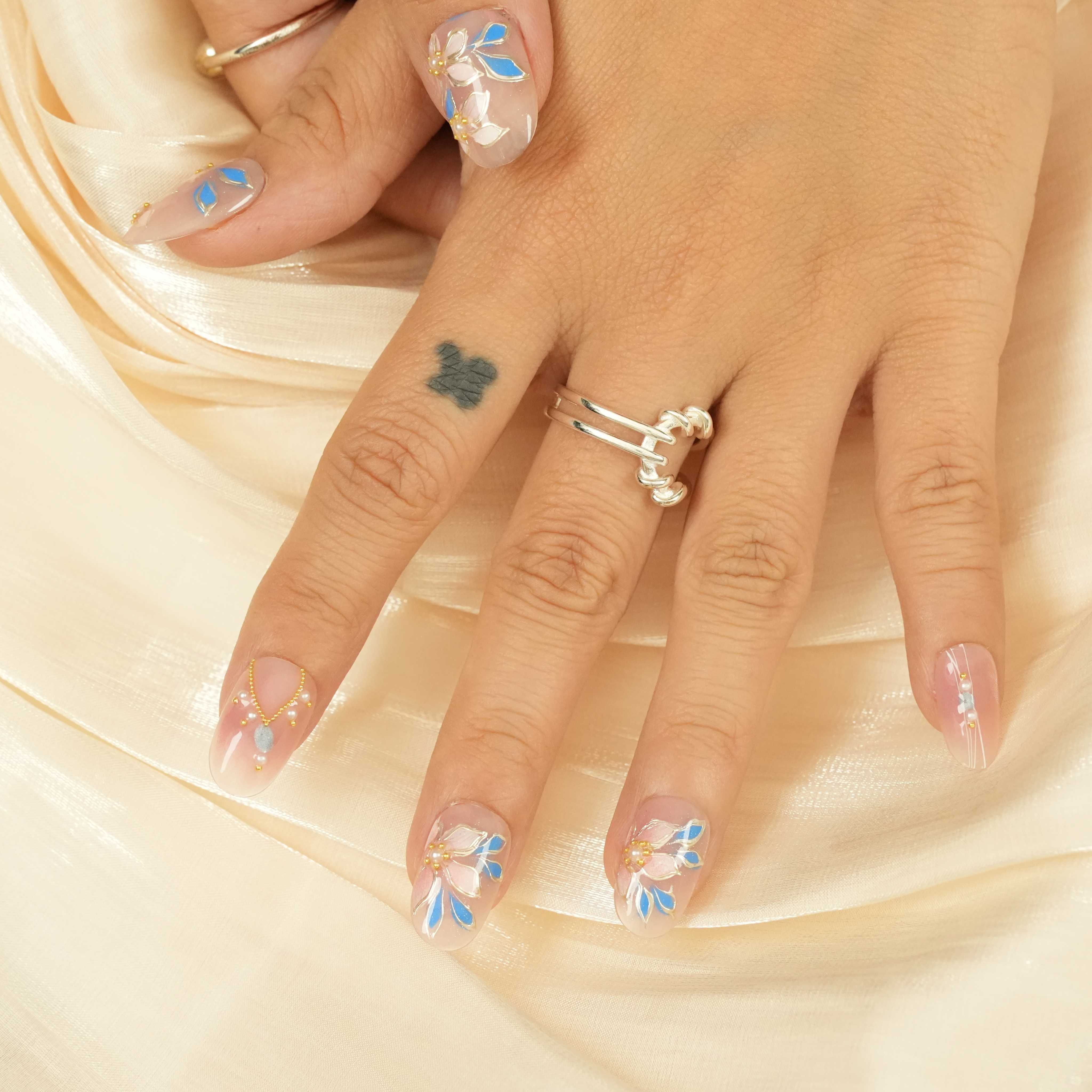 Short floral stick on nails laid out on soft fabric, showcasing delicate blue and white hand-painted flowers and gold details