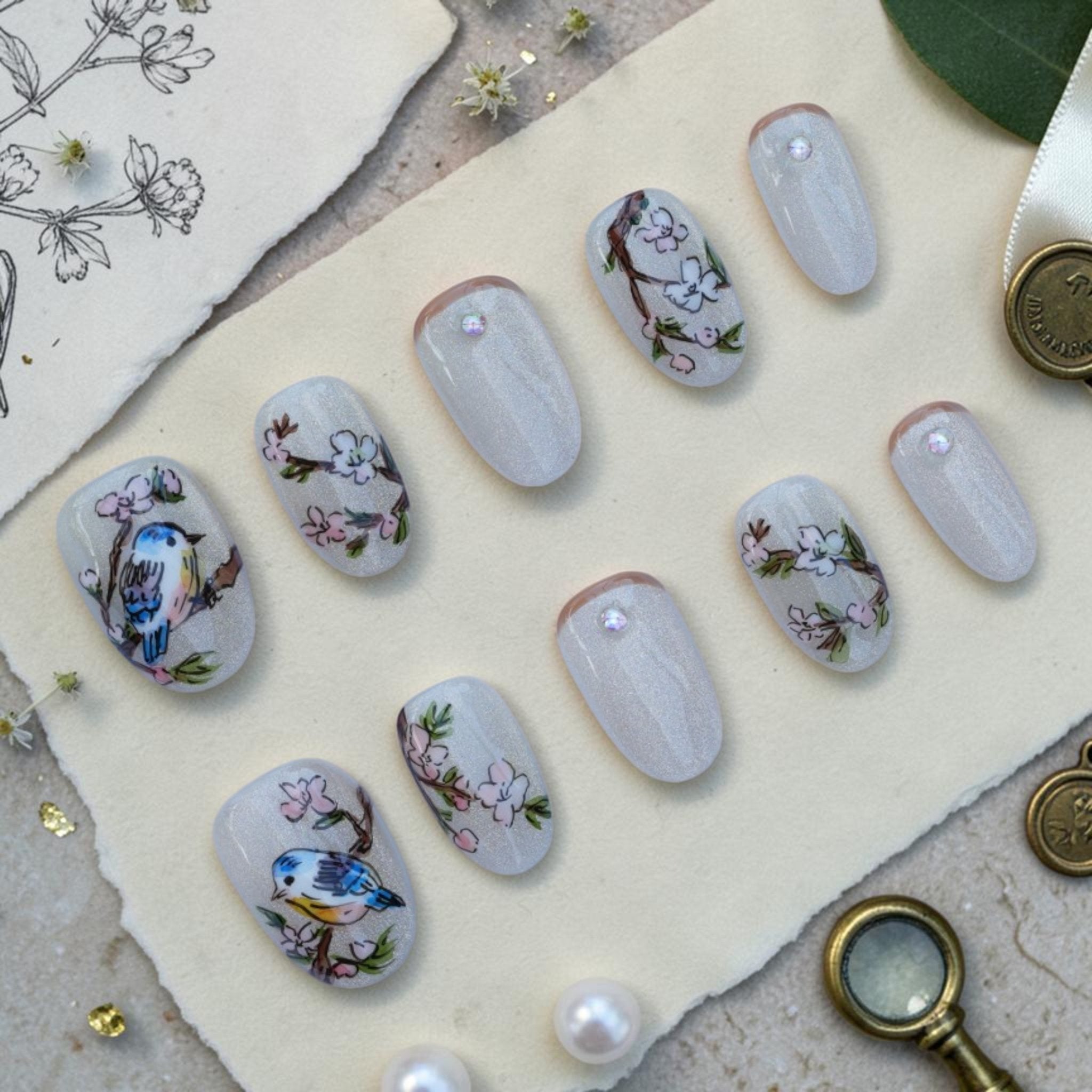 Handmade bird press on nails flat lay with hand painted songbirds and blossoms on a pearl cat eye base