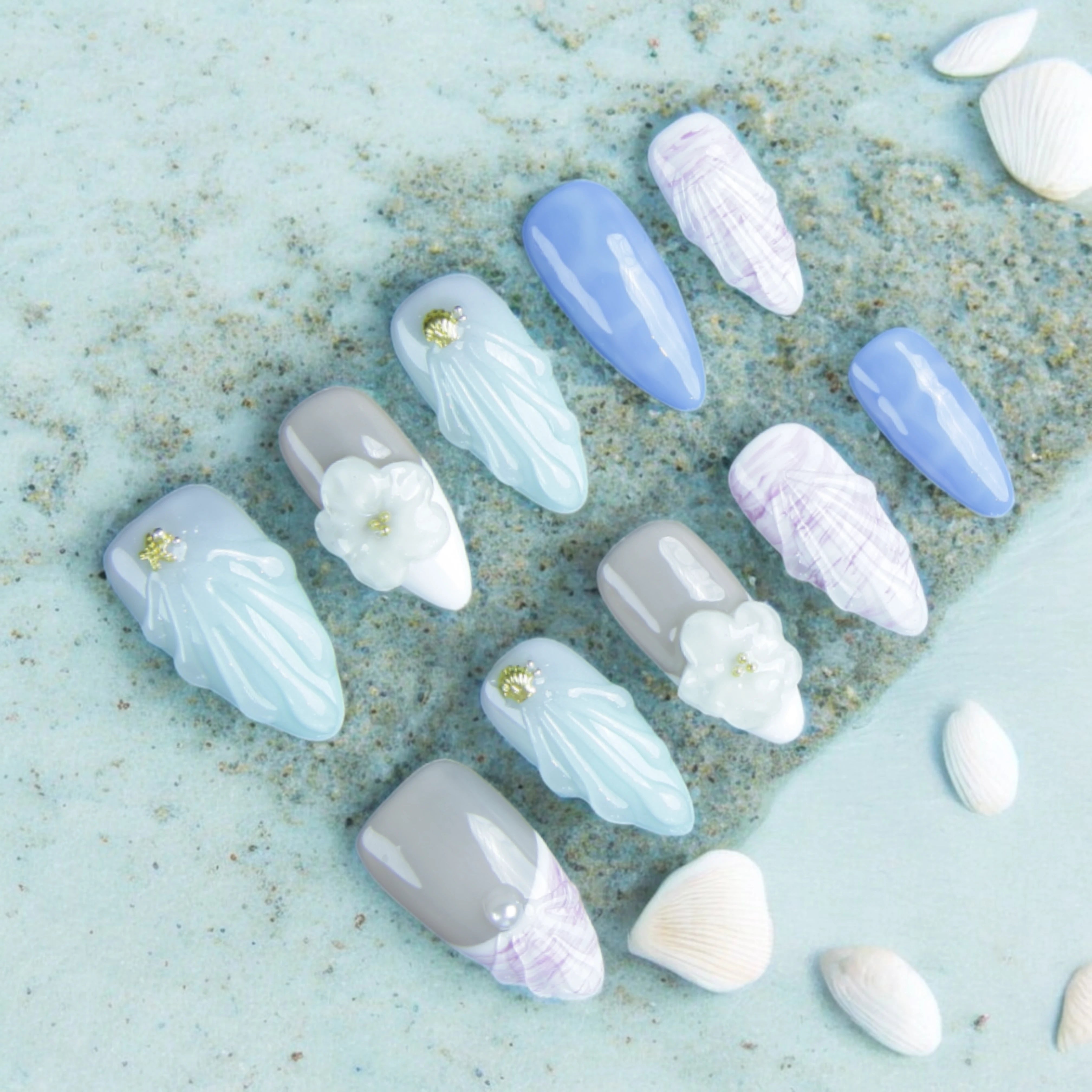 A set of Mystic Ocean press on nails displayed on a soft blue sandy background, featuring delicate shell textures, floral accents, and oceanic hues.