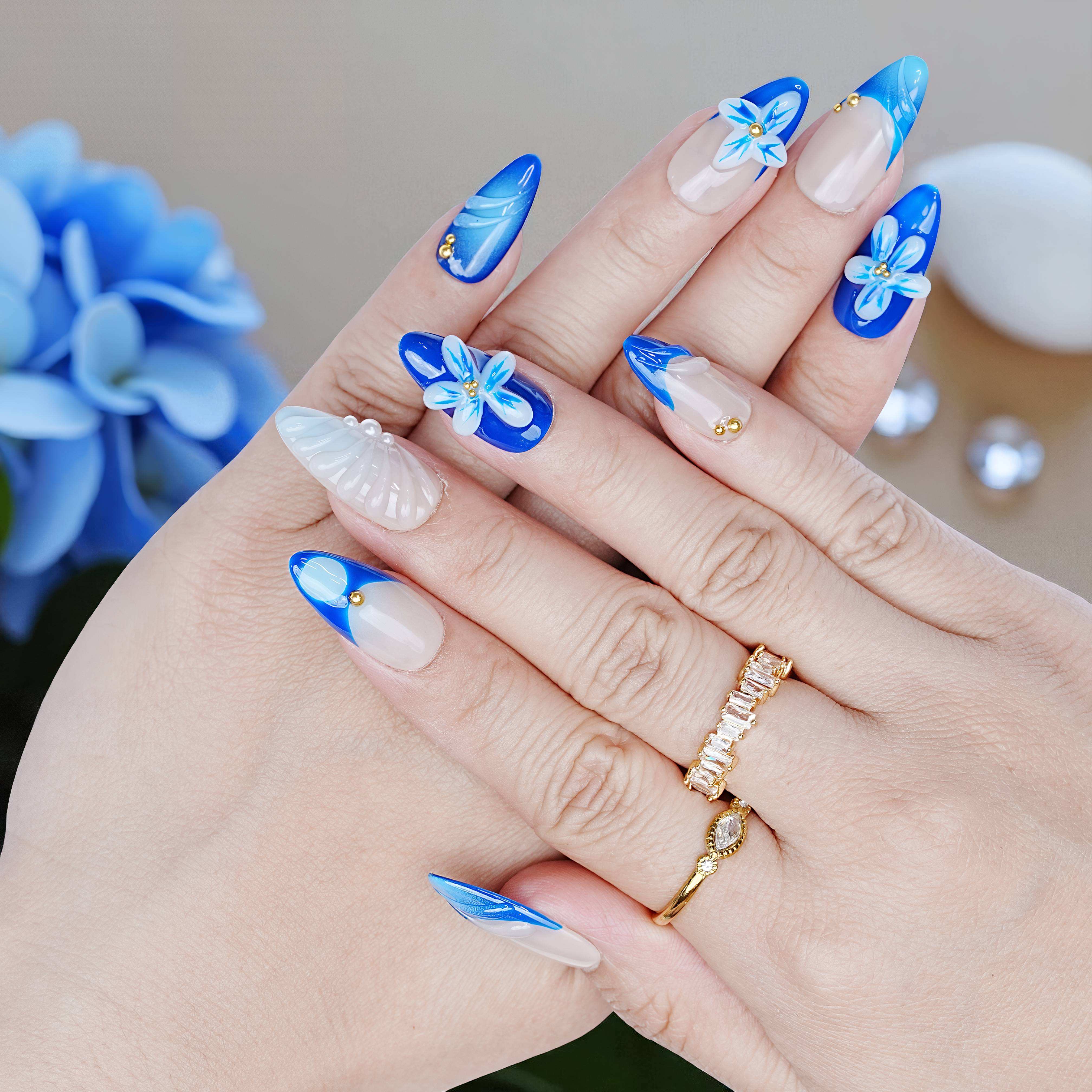 Hand pose showing full Blue Mirage nail set with deep blue tips, floral details, and glossy finish, surrounded by blue decor.