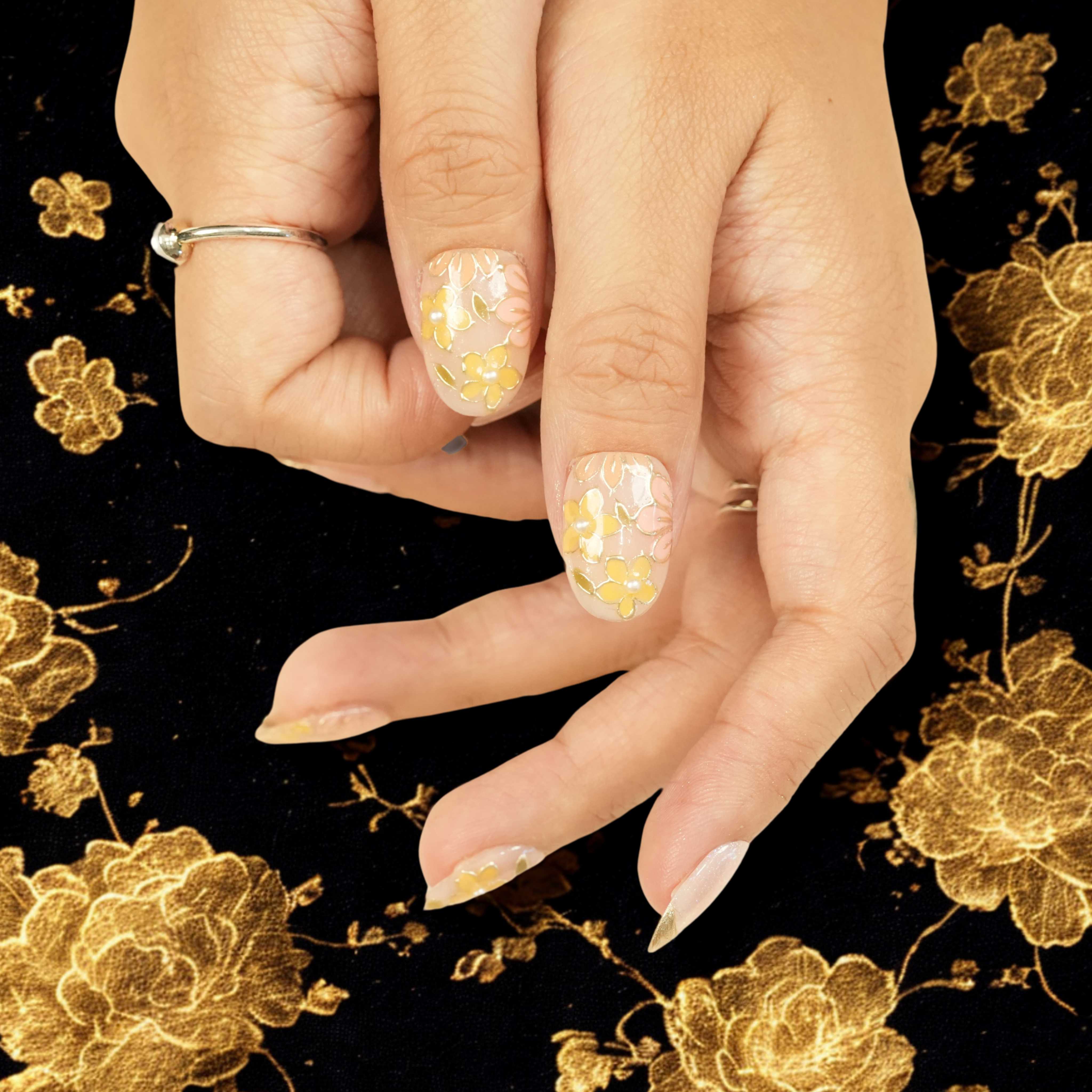 False nail with golden floral design and shimmer finish, shown on thumb against black floral background. Stick on nail design