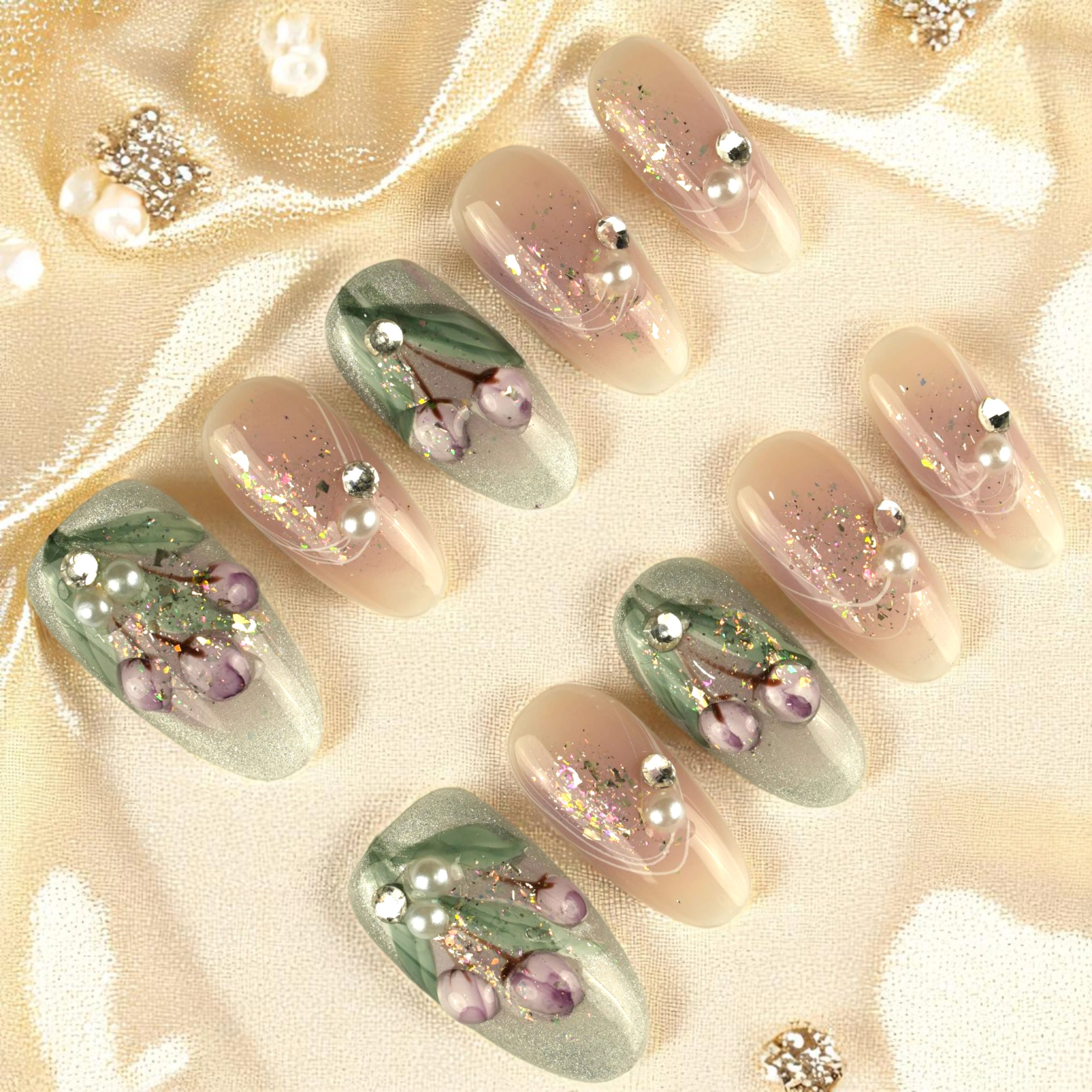 Flat lay of press on nails on satin background, combining nude and sage green tones with purple flowers, glitter and pearl accents