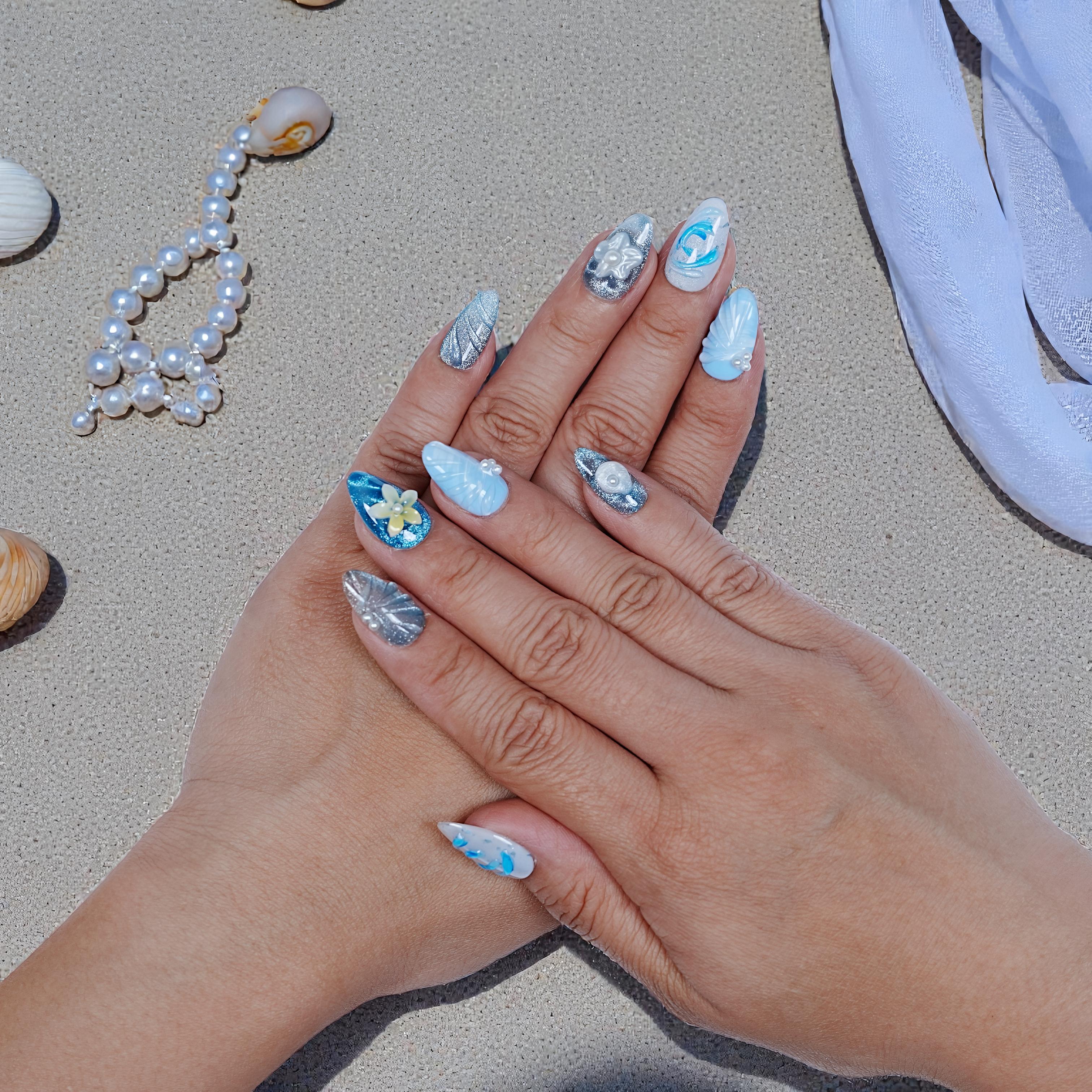 Hand with beach-themed blue and silver handmade press on nails, pearl and flower accents on sandy surface