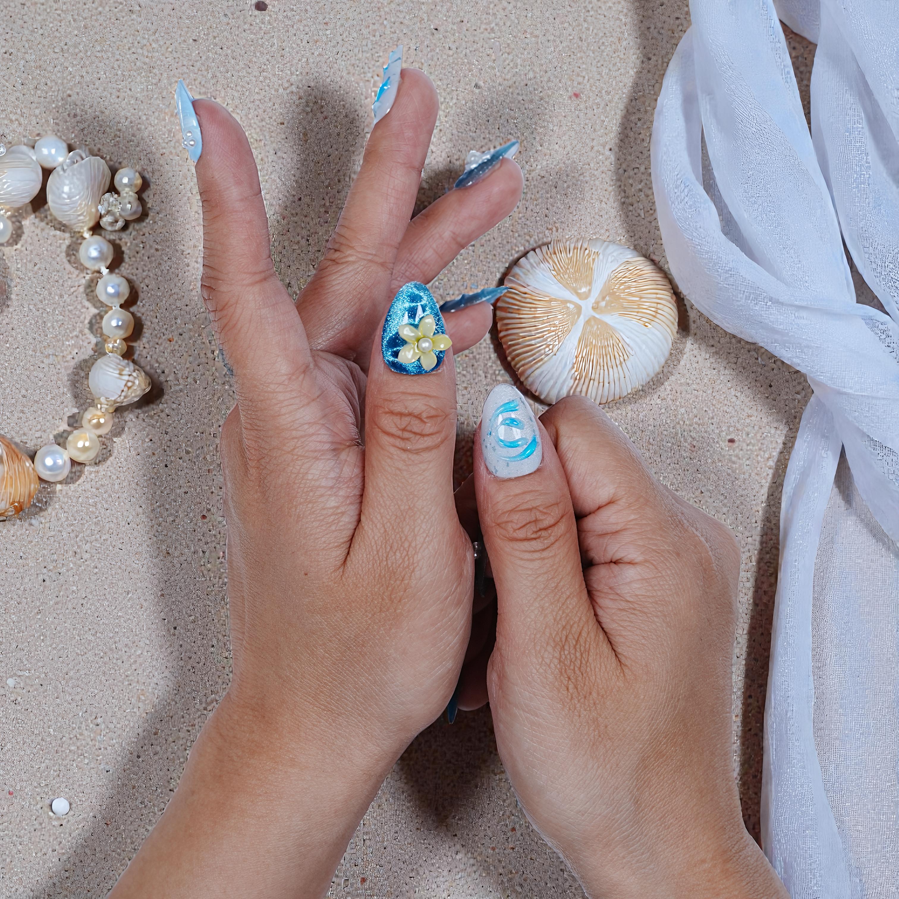 Hands with premium handmade blue and white press on nails featuring pearl flower design on sandy beach background with seashells and white sheer fabric