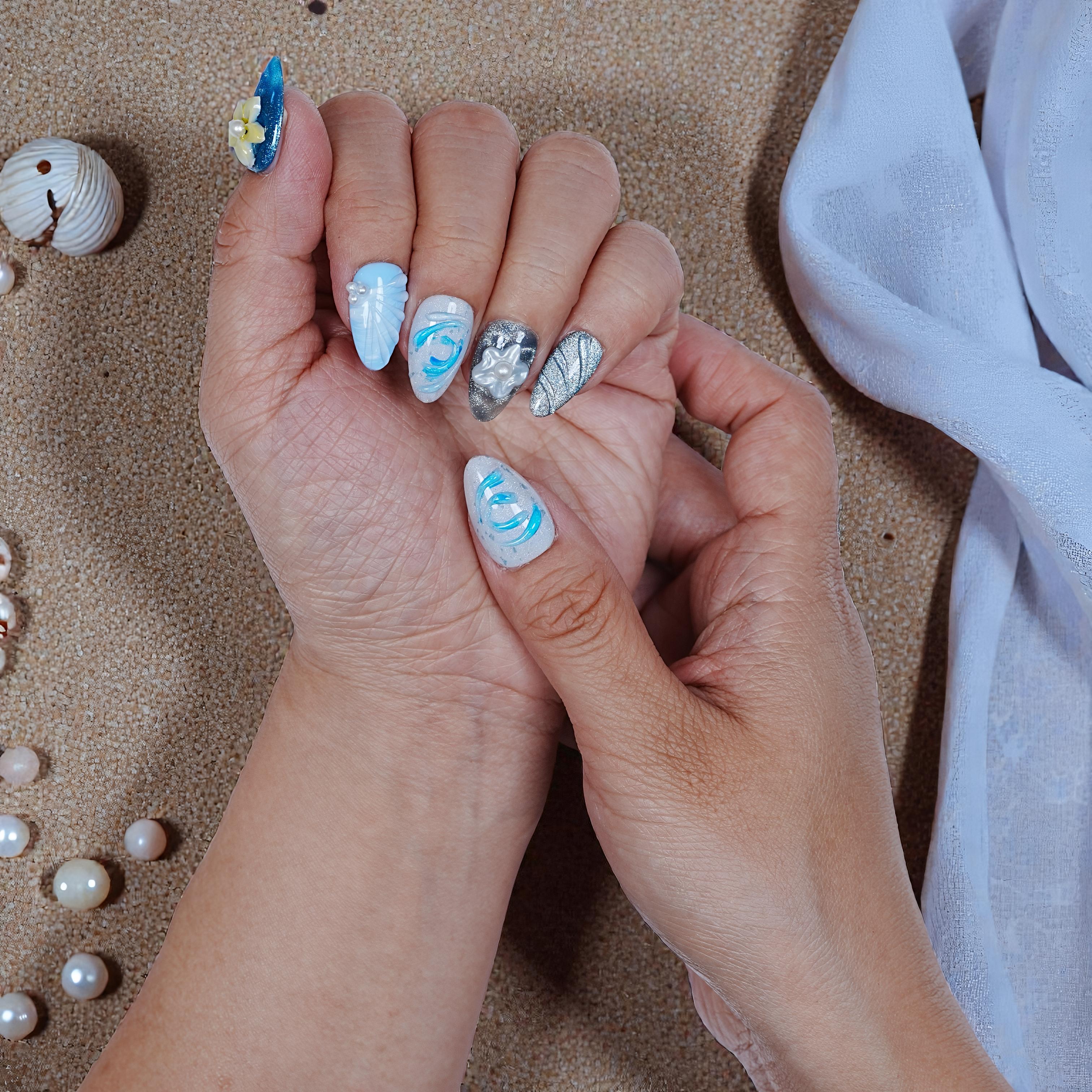 Hand with blue and silver handmade press on nails featuring floral and wave designs on sandy background