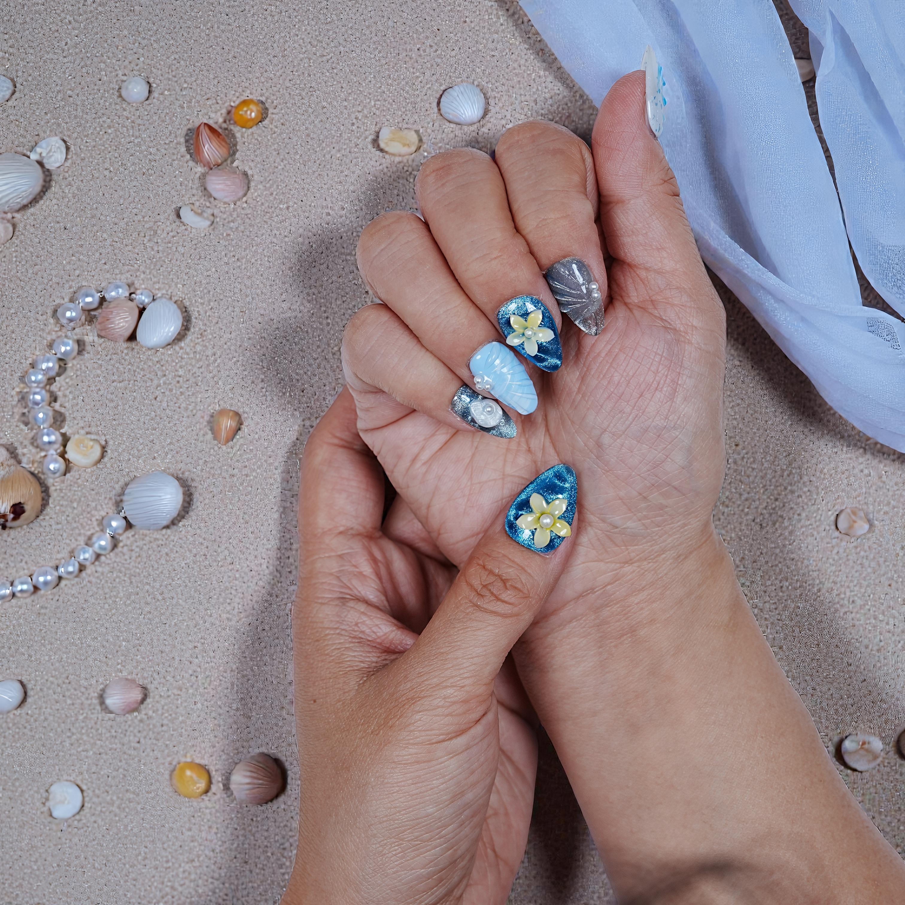 Close-up of hands with premium handmade press on nails featuring blue and silver designs with flowers and pearls on sandy background