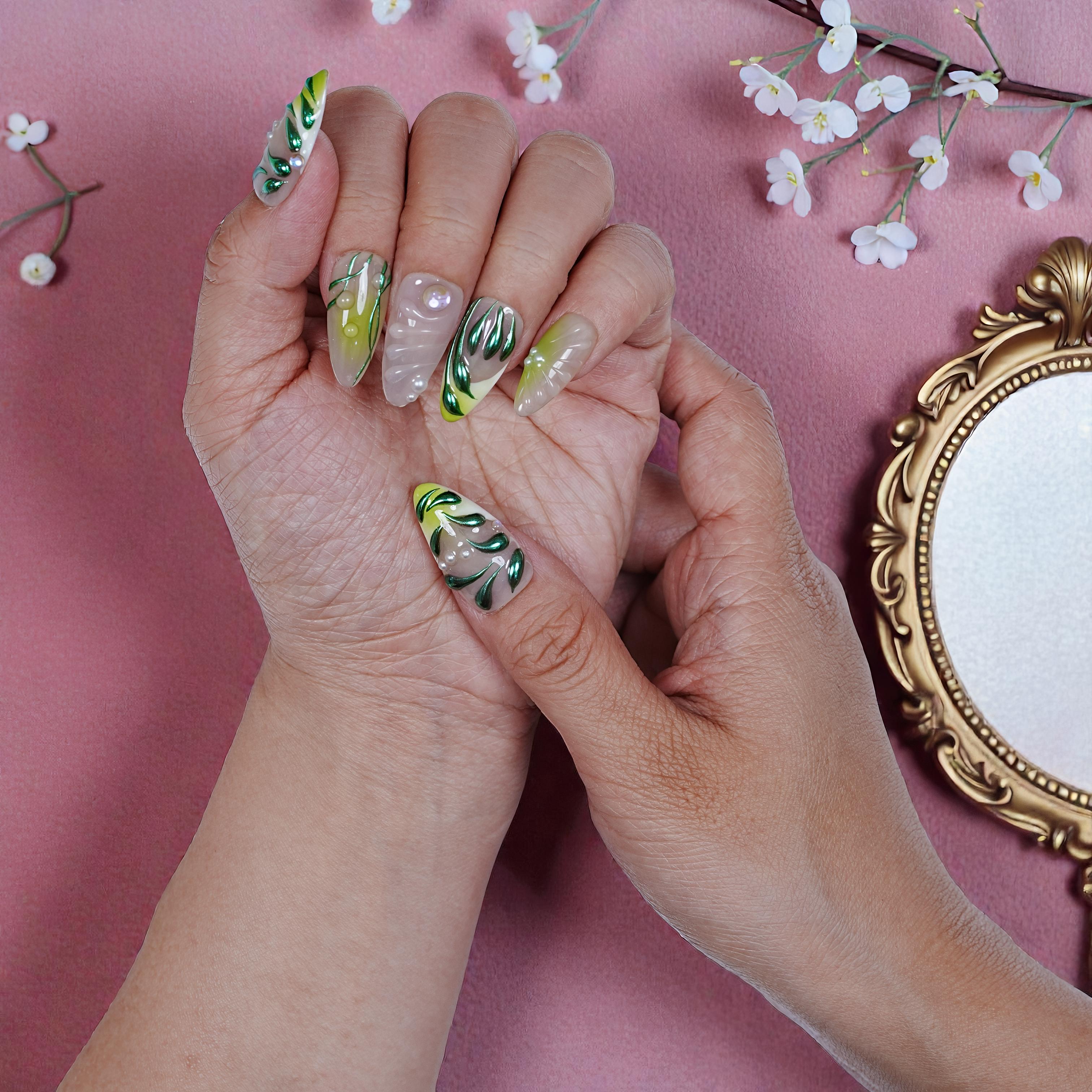 Hand with long, handmade press on nails featuring green leaf designs and pearls on a pink background with flowers and gold-framed mirror