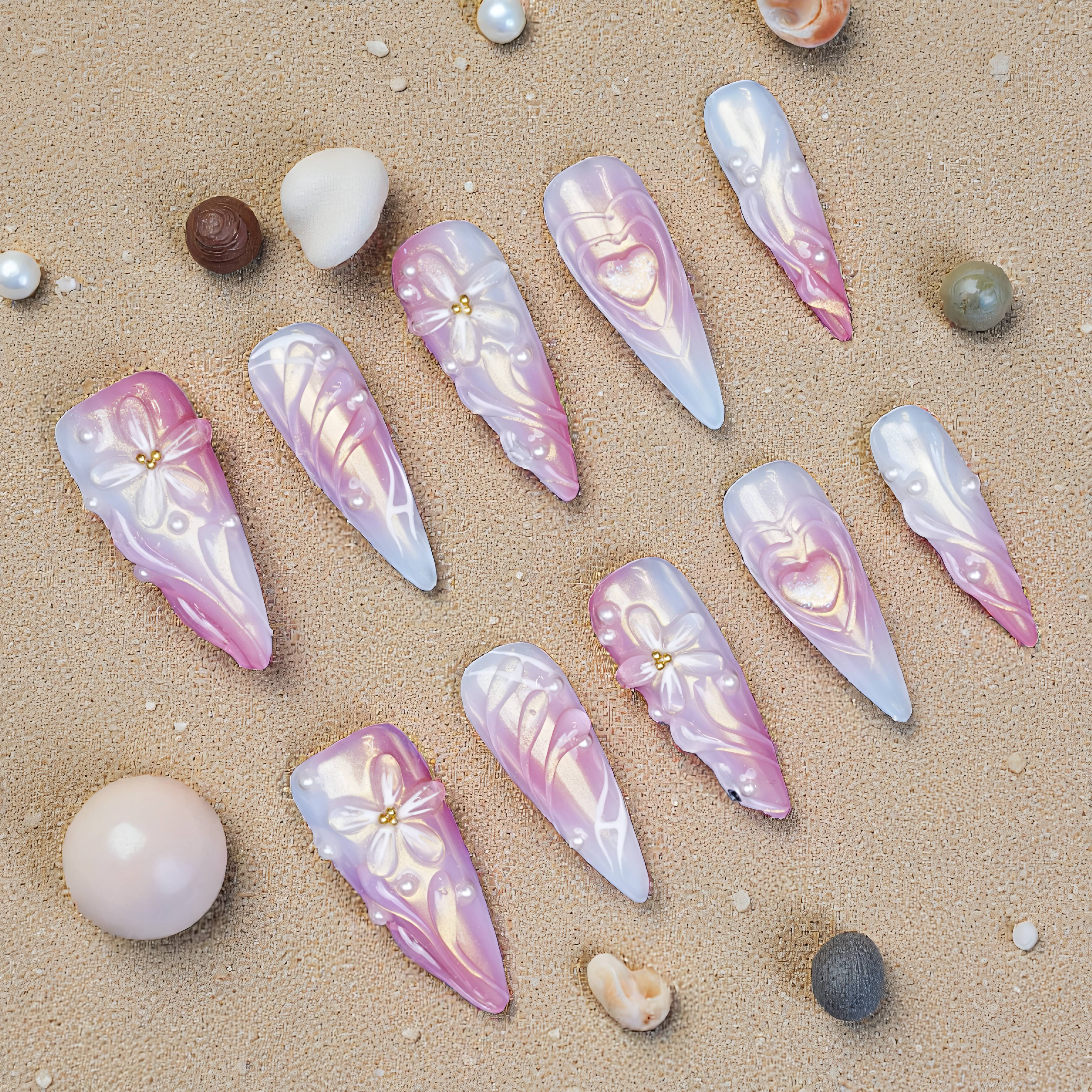 Long stiletto press on nails in pink and white chrome with 3D bows, hearts, and pearl accents displayed on sand with shells