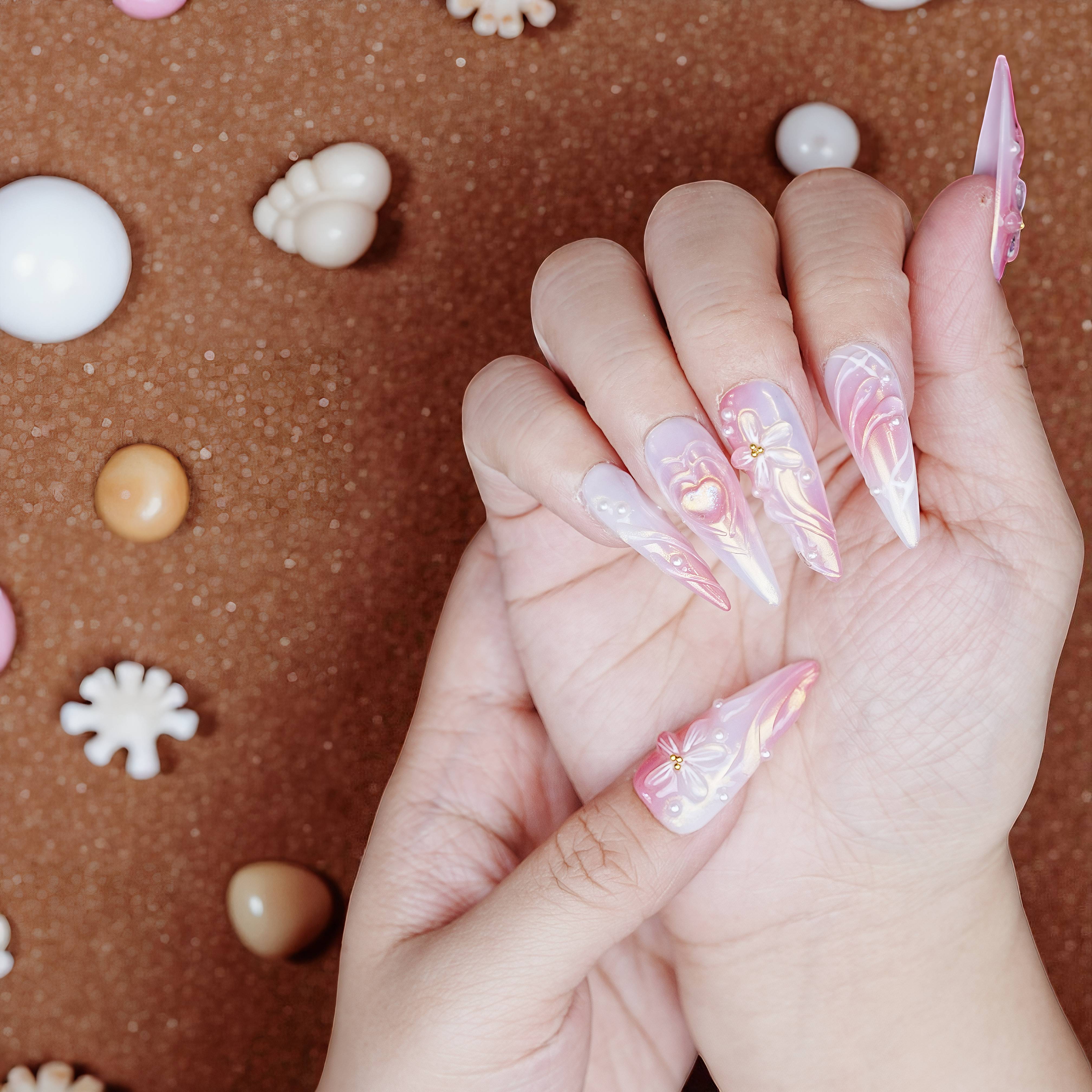 Hand view of pink chrome heart nails with bow designs and pearly white stiletto tips surrounded by seashell props