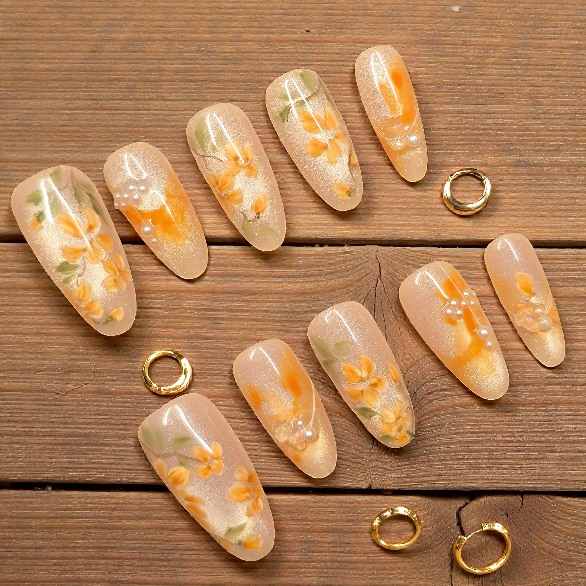Handmade golden shimmer floral nails with orange petals, pearl accents, and soft glossy finish, reusable and reshaped easily with nail file.