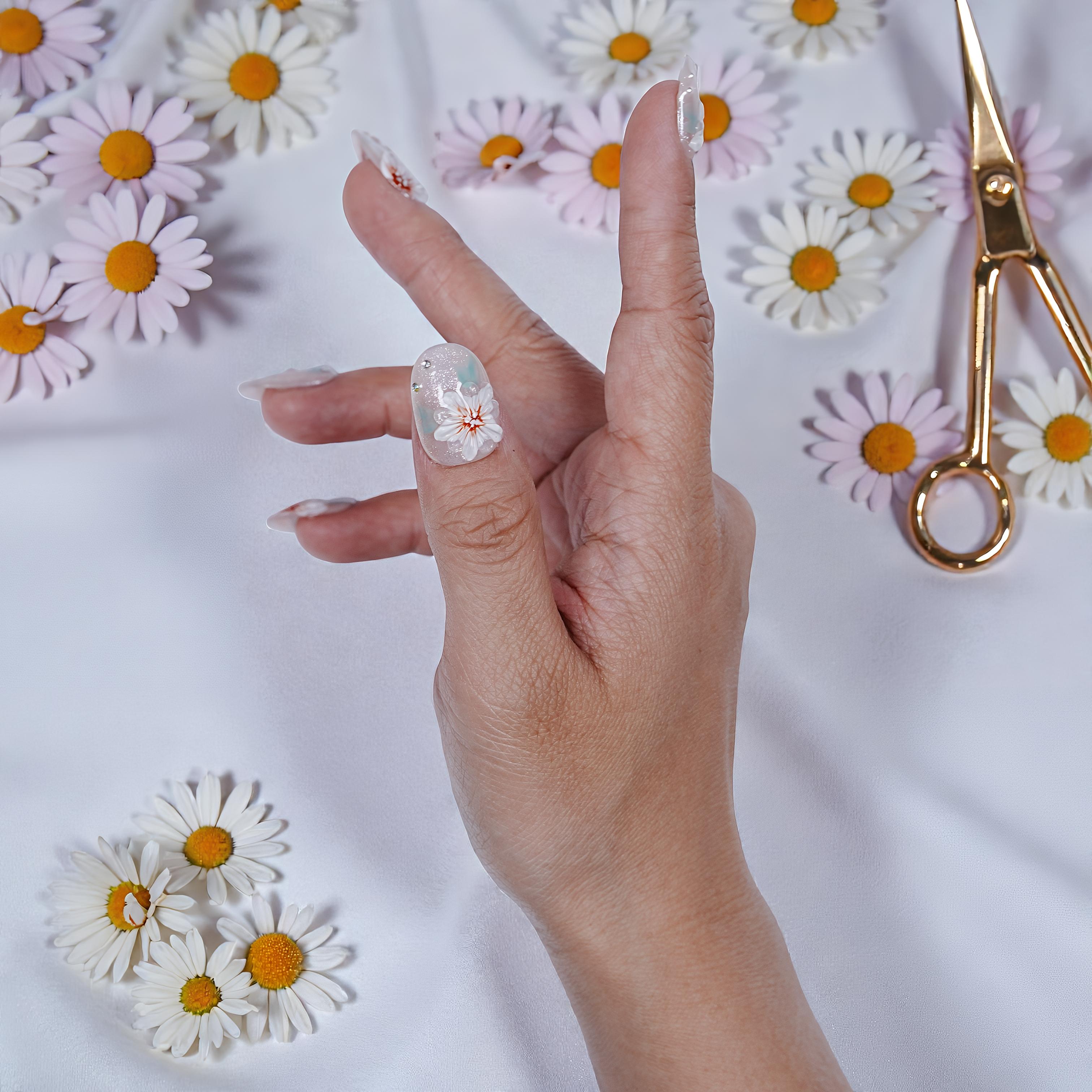 Hand with clear press on nails featuring white floral designs, surrounded by daisies on white fabric