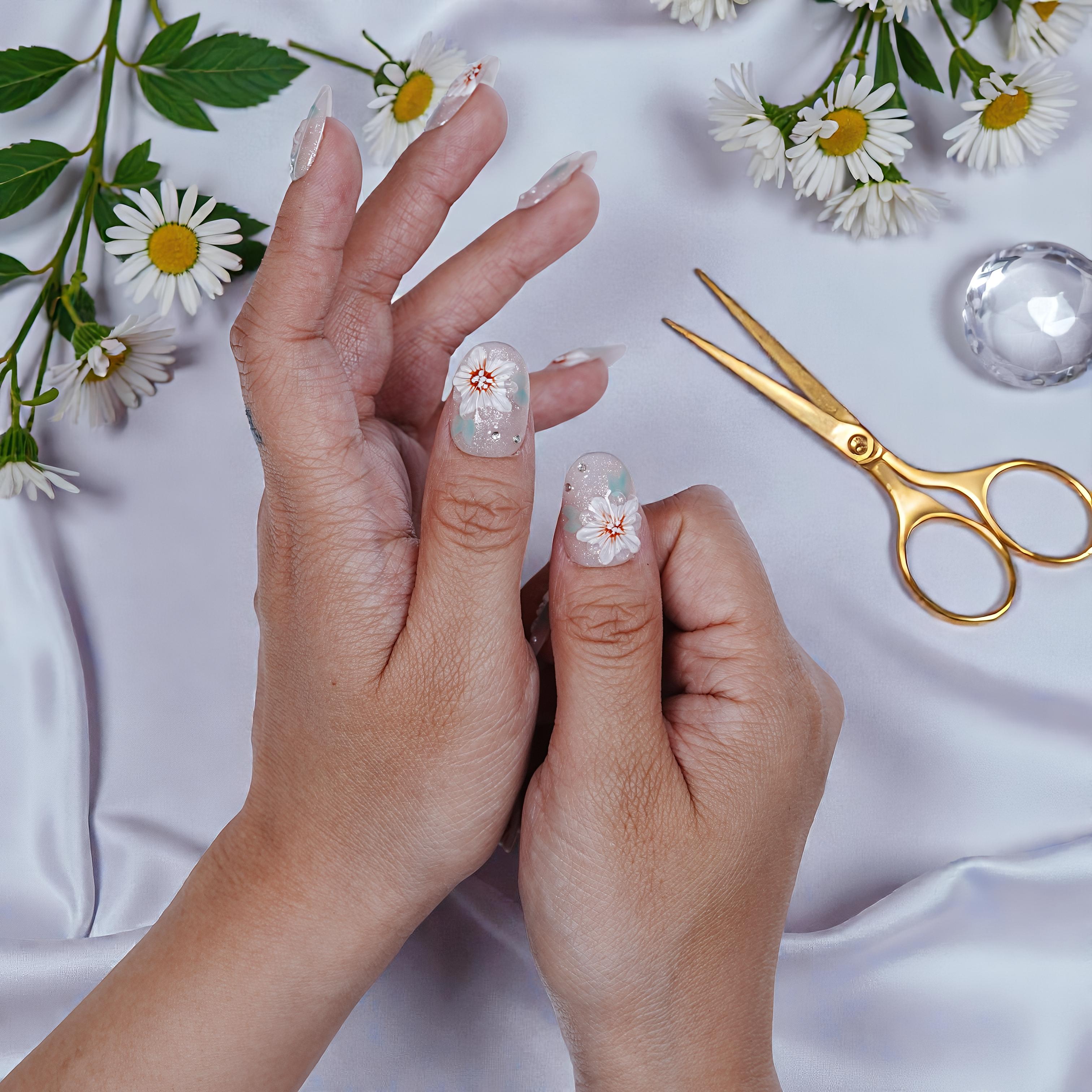 Hand with transparent press on nails featuring white floral design, surrounded by daisies and gold scissors on silk
