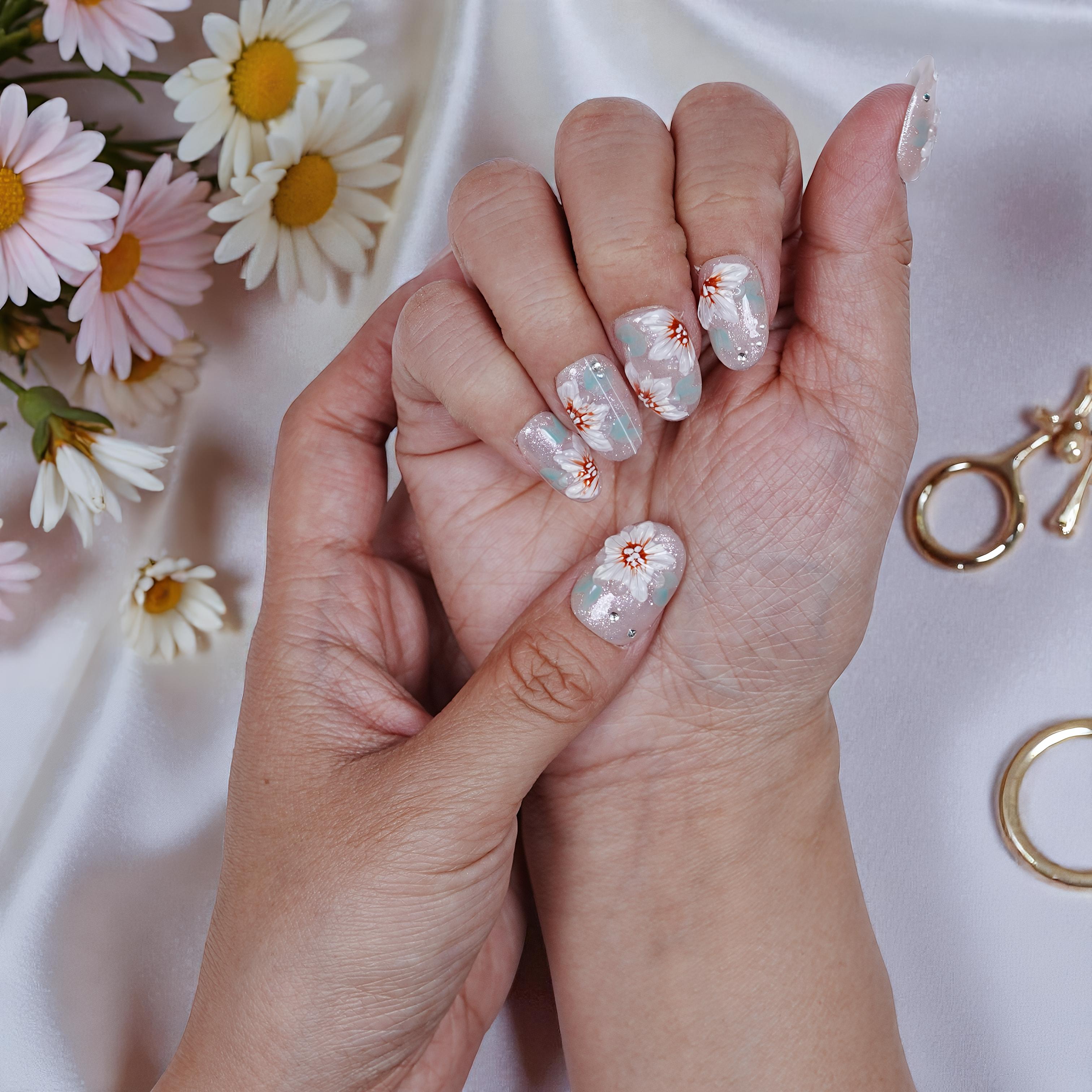 Hands showing reusable press on nails with white floral design on glittery pink base surrounded by daisies and gold rings