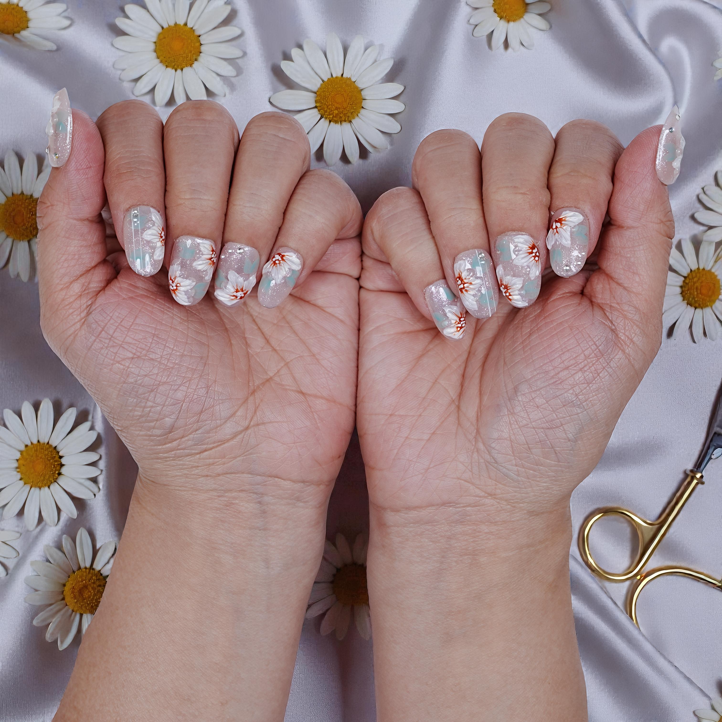 Hands with premium handmade press on nails featuring white and orange floral design on glittery background, daisy flowers and gold nail scissors on satin