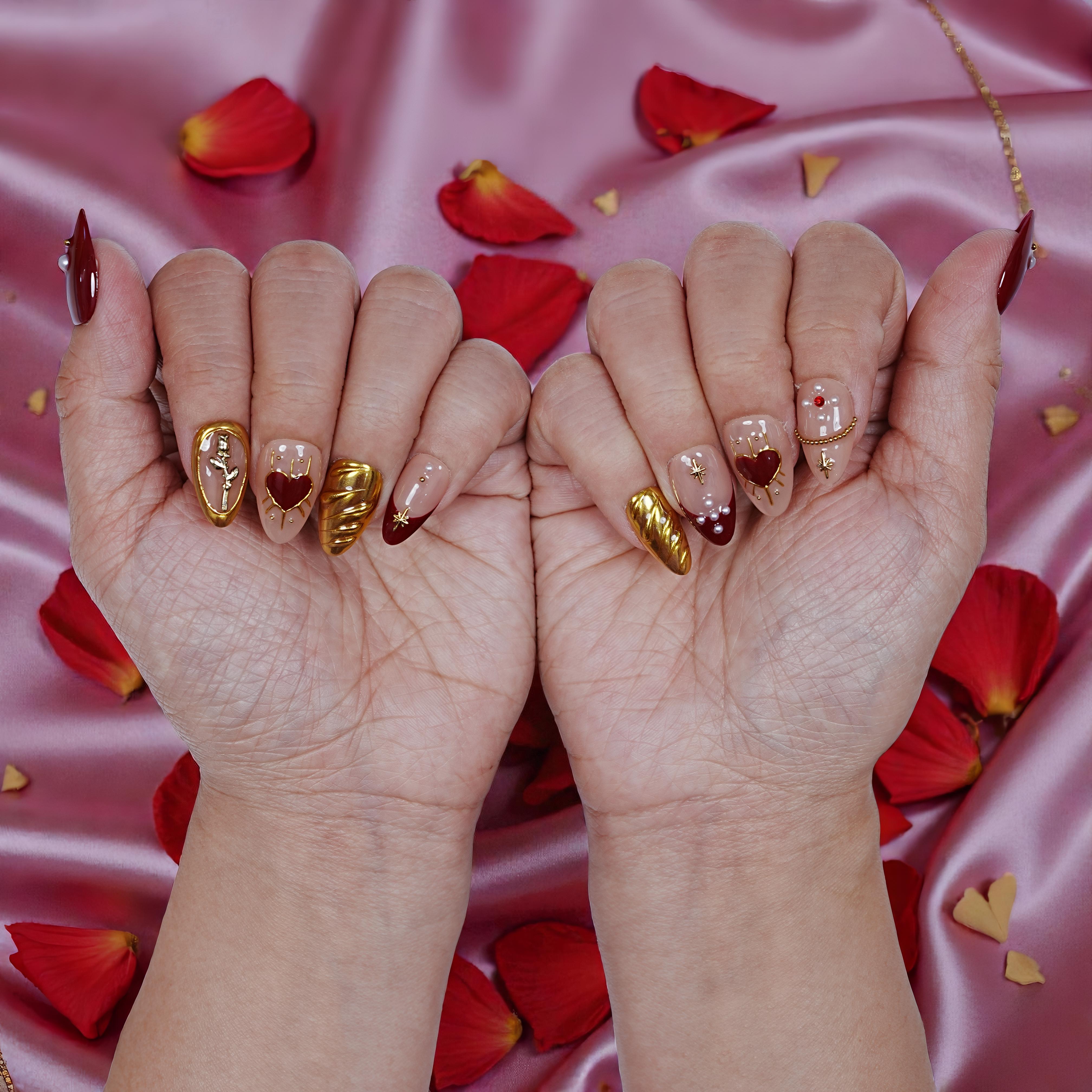 Handmade press on nails with gold, red, and heart designs on hands, rose petals on pink silk