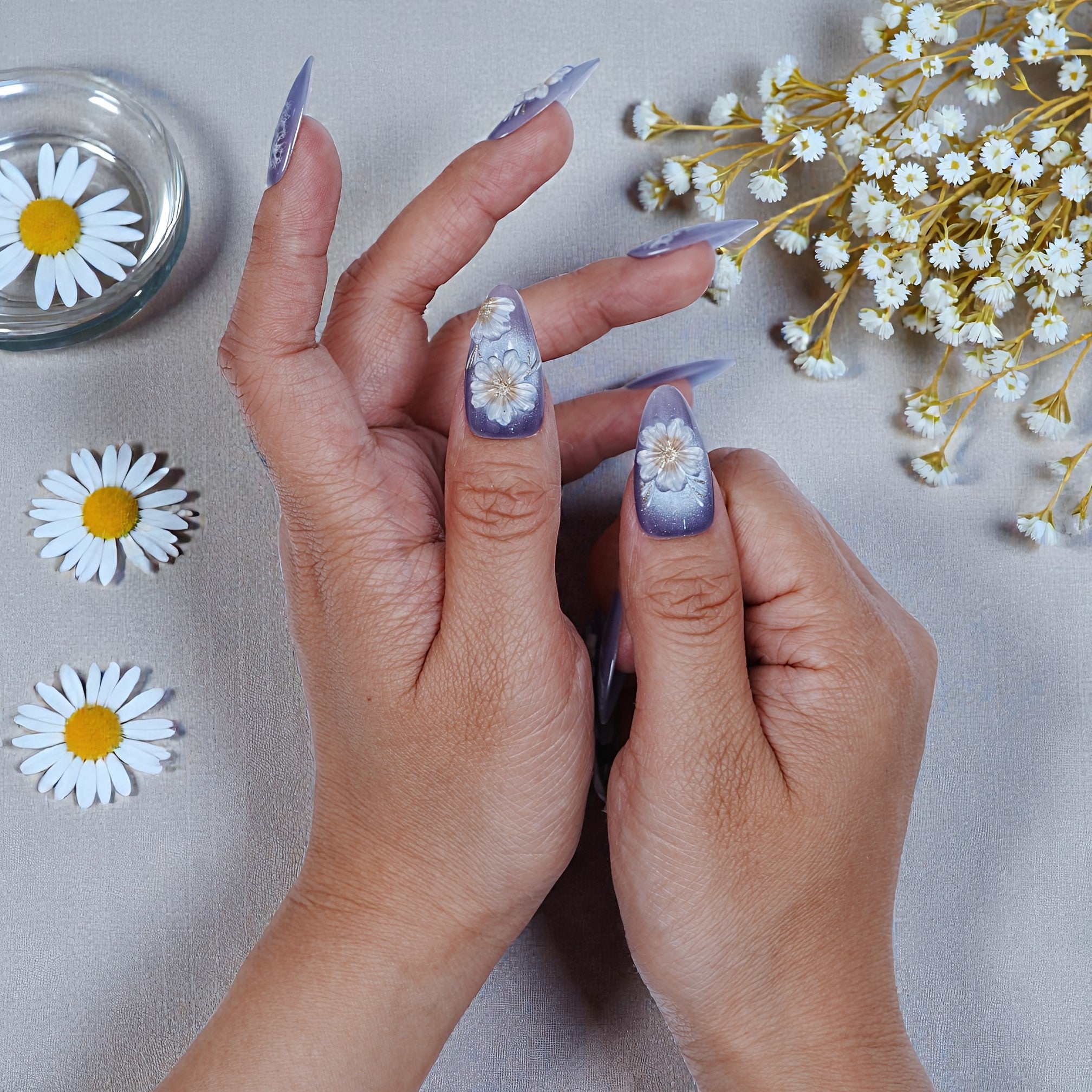 Hand with purple ombre press on nails featuring white daisy designs, surrounded by daisies.