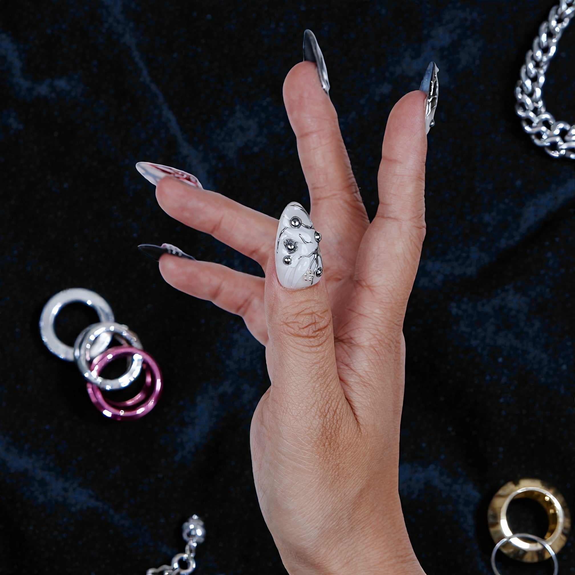 Hand with premium handmade press on nails featuring silver marble and bead accents on black background, surrounded by metallic jewelry.