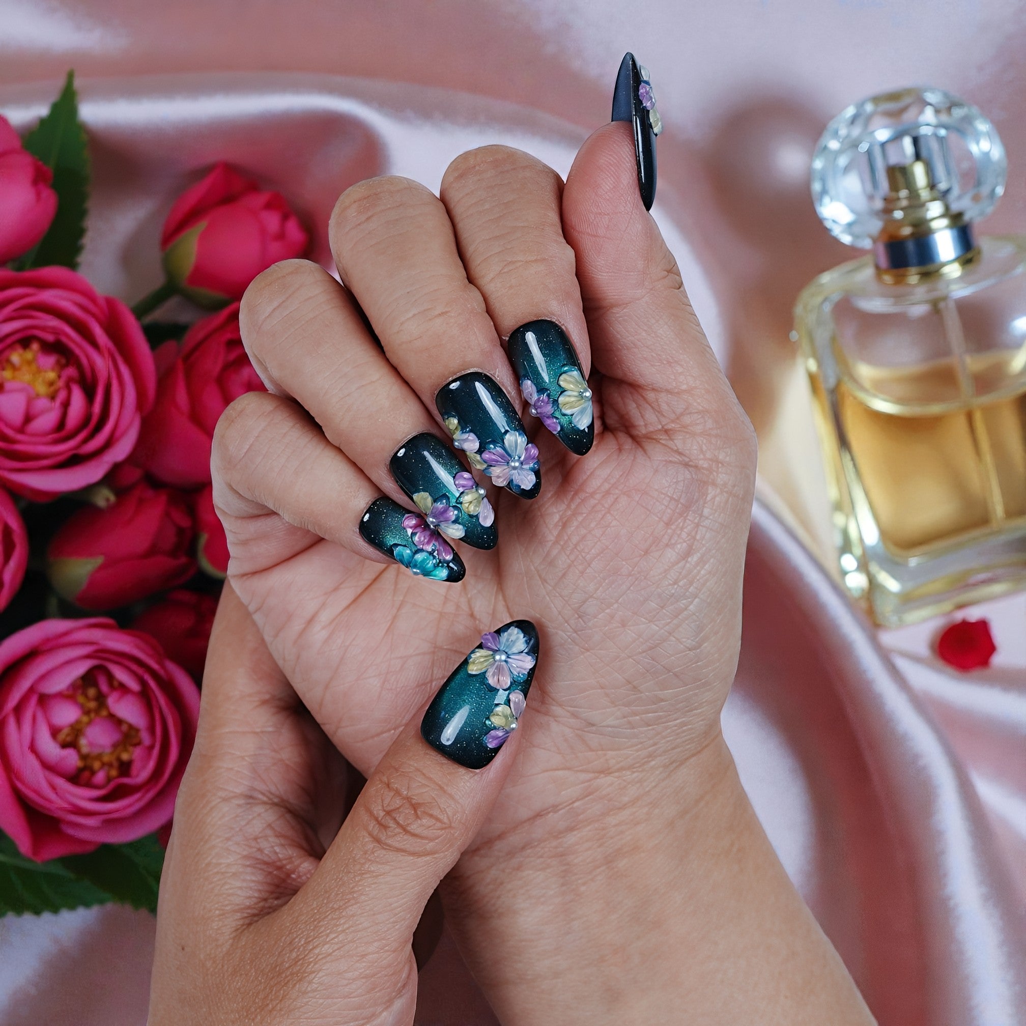 Hand with dark floral press on nails, roses, and perfume on pink satin background