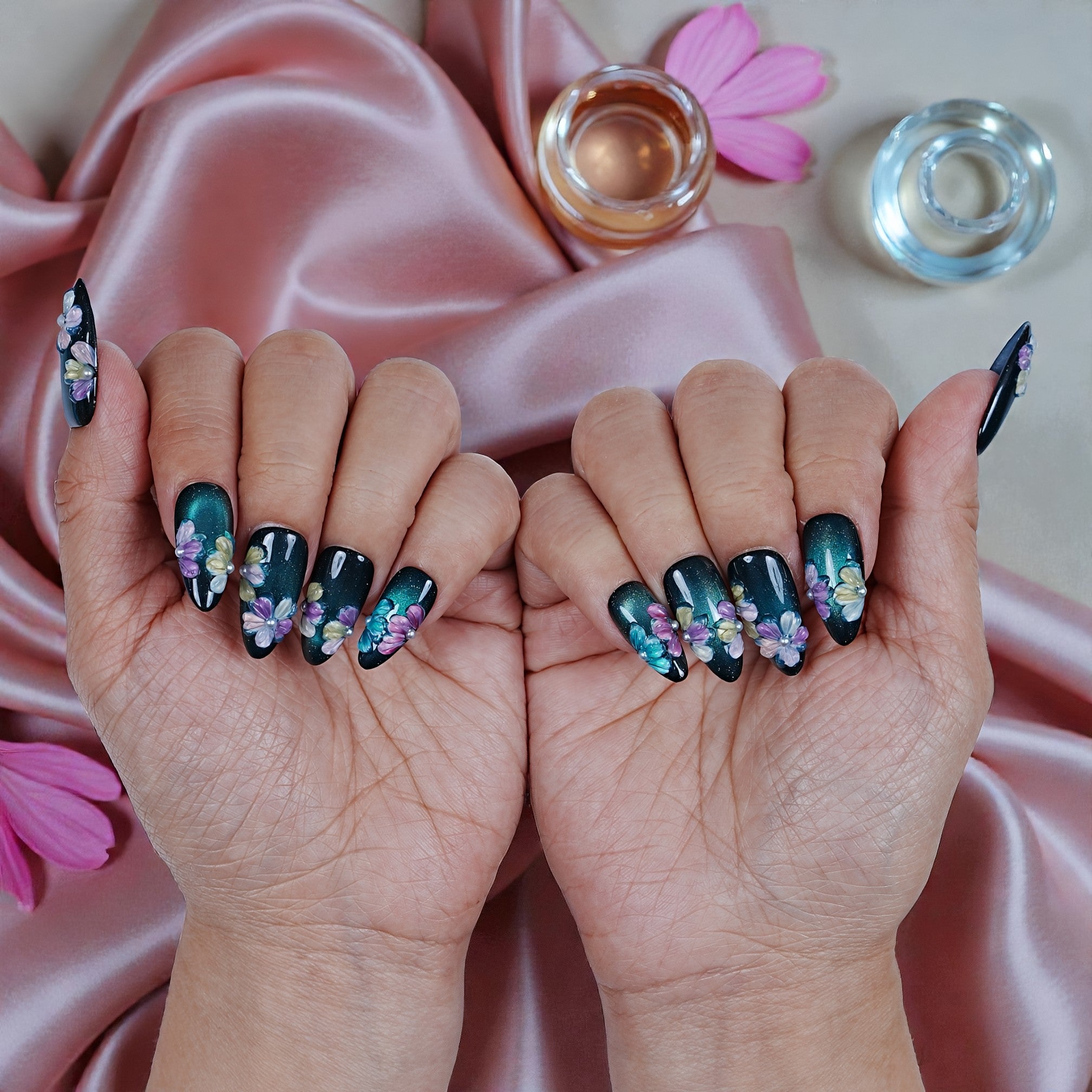 Handmade floral press on nails in dark green, displayed on hands over pink satin fabric.
