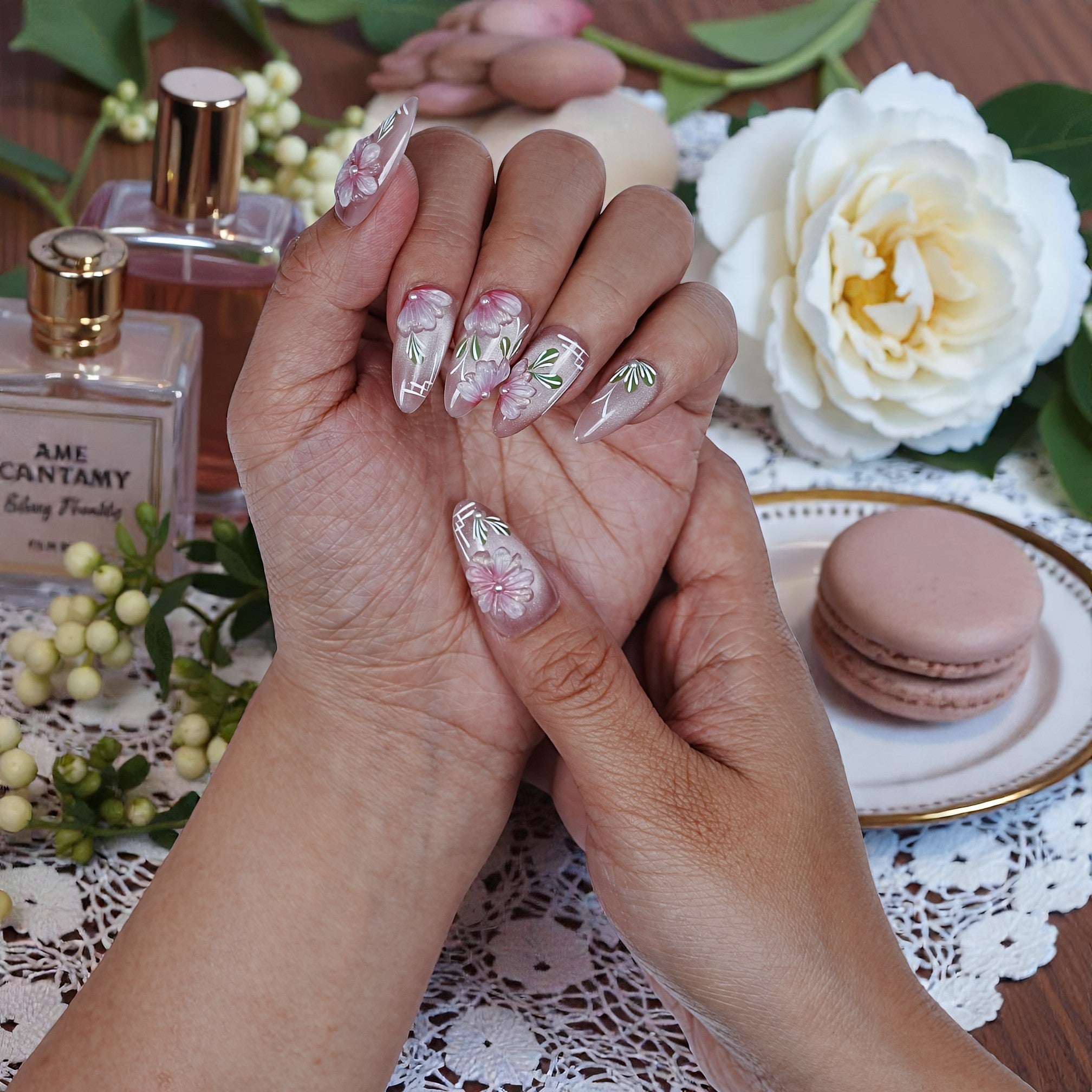 Hand with premium handmade press on nails featuring pink floral design, surrounded by perfume, flowers, and a macaron on a lace tablecloth.