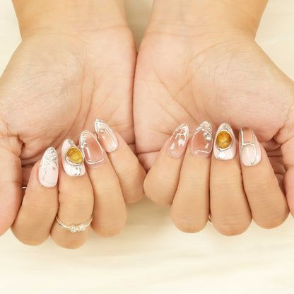 Hands with almond-shaped press on nails featuring nude shades, silver metal details, and gold amber accents