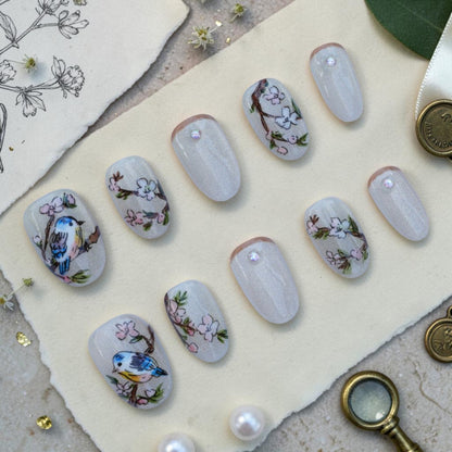 Handmade bird press on nails flat lay with hand painted songbirds and blossoms on a pearl cat eye base
