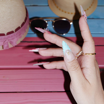 Side angle hand pose showing gradient press on nails with beachy pink hues and floral decals, hat and sunnies in background