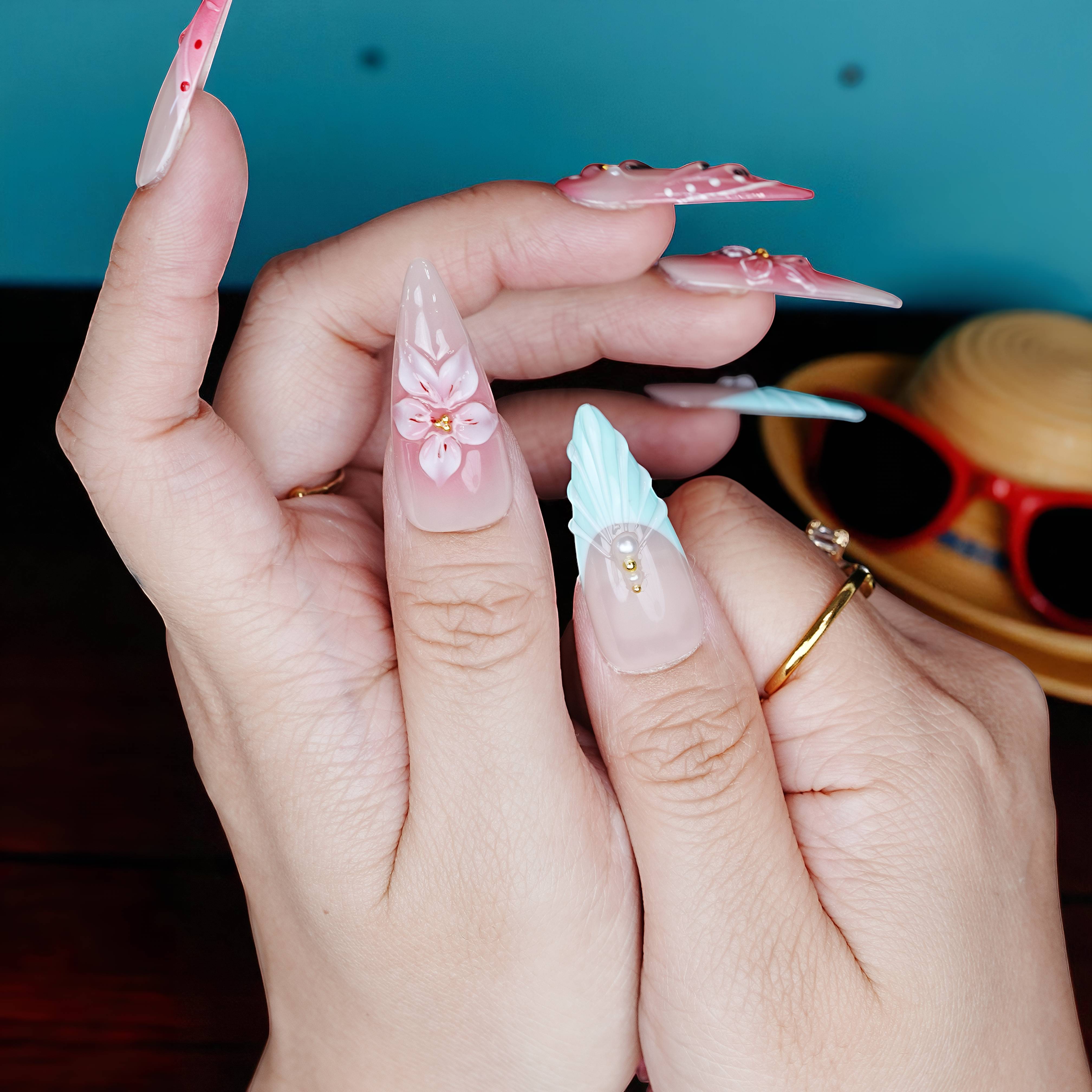 Hands displaying long stiletto press on nails with alternating aqua tips and pink floral patterns under moody lighting