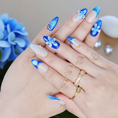 Hand pose showing full Blue Mirage nail set with deep blue tips, floral details, and glossy finish, surrounded by blue decor.