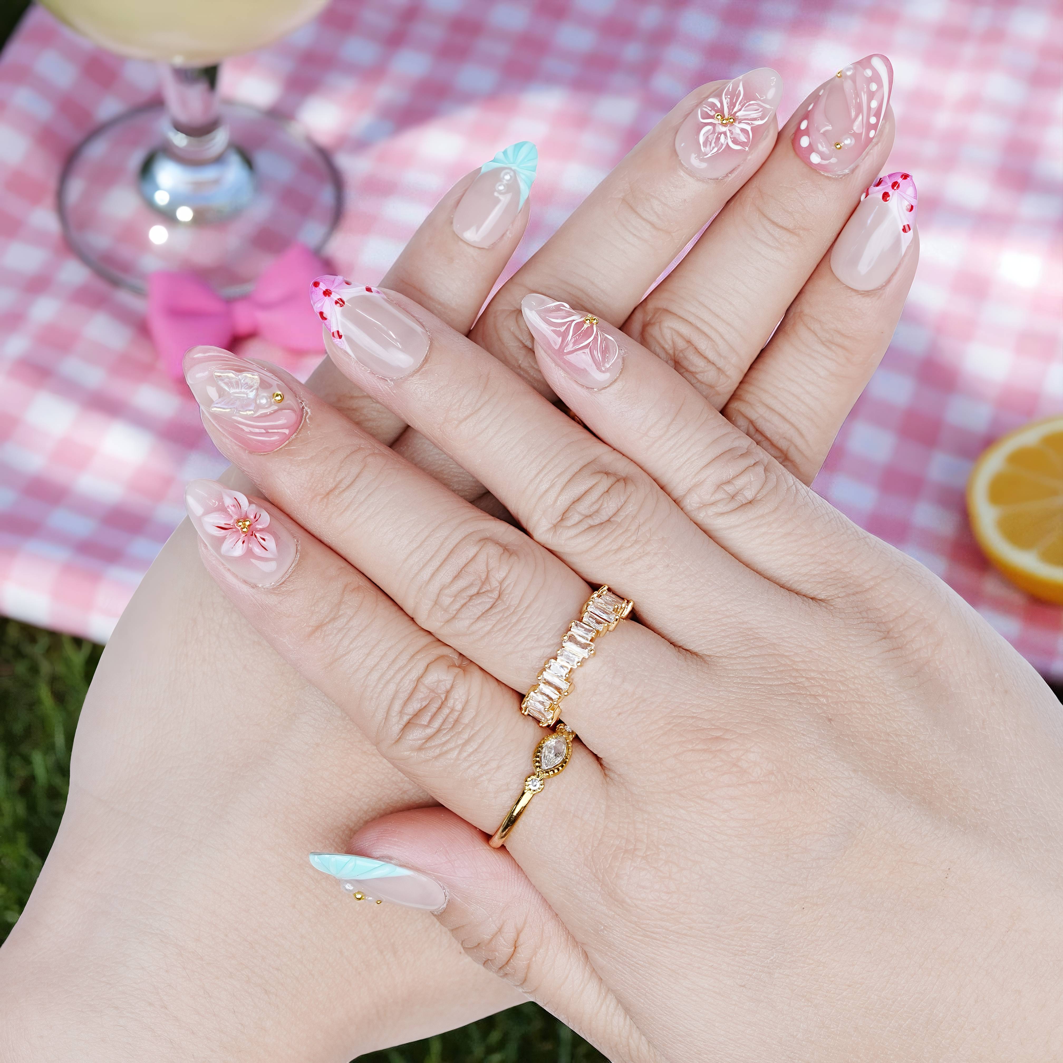 Hand pose showing Peachy Petal nails with floral and pink swirl details, styled with soft drinks and gold rings.