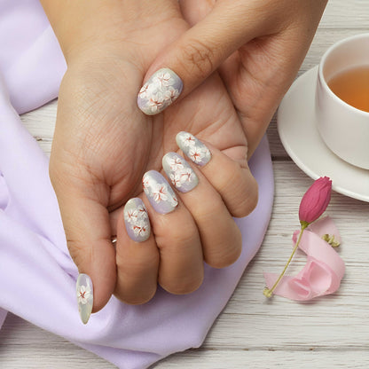 Close-up of hands with floral nail design, wearing a light purple sleeve, with a cup of tea and pink tulip on a wooden surface.