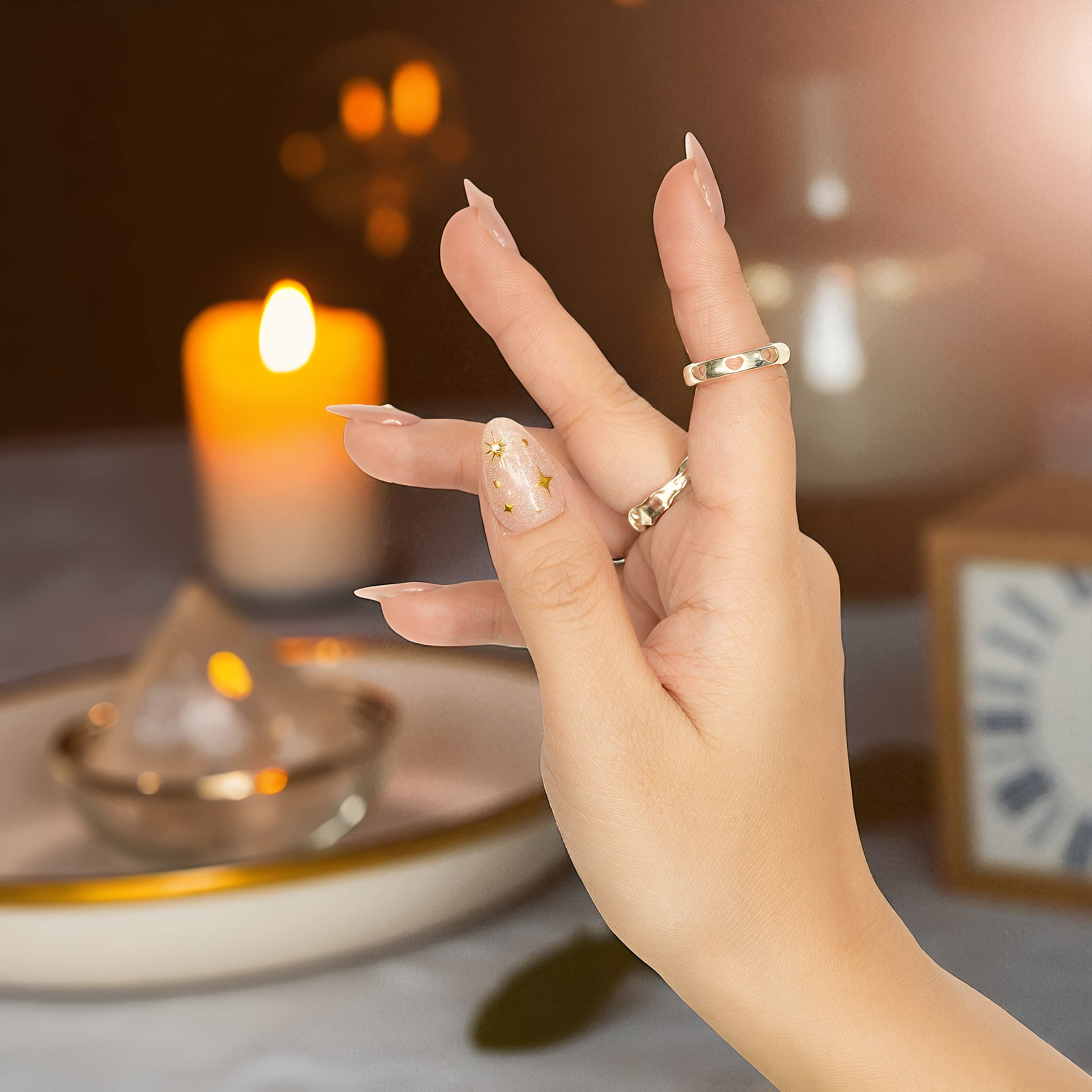 Hand posed against soft background showing false nails in nude base, featuring delicate gold foil and crystals.

