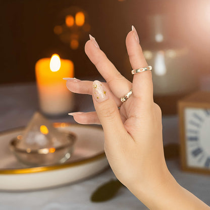 Hand posed against soft background showing false nails in nude base, featuring delicate gold foil and crystals.

