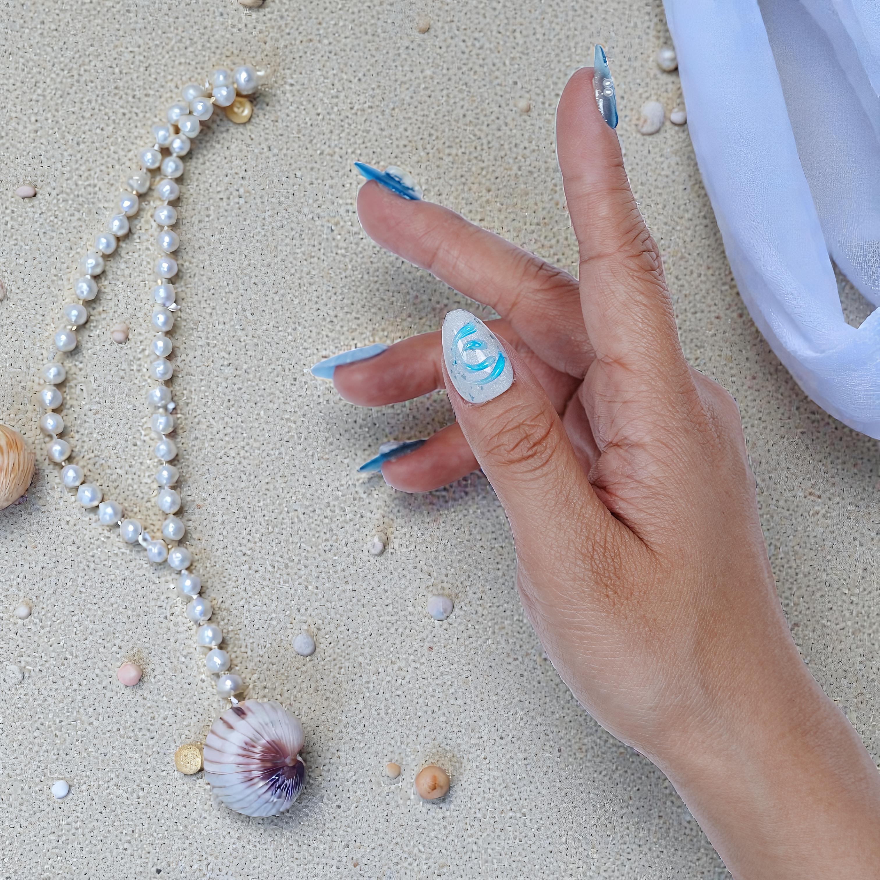 hand with long blue and white handmade press on nails on textured sandy surface with pearl necklace and seashells