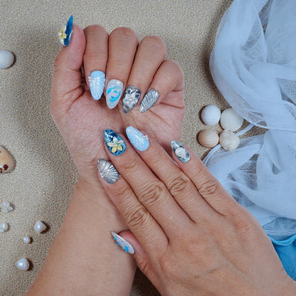 Hands with blue, silver, and white handmade press on nails featuring pearls and floral 3D designs on sandy background