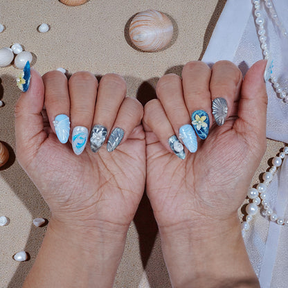 Close-up of hands with premium handmade press on nails in ocean blue and silver, decorated with pearls and floral accents on sandy background
