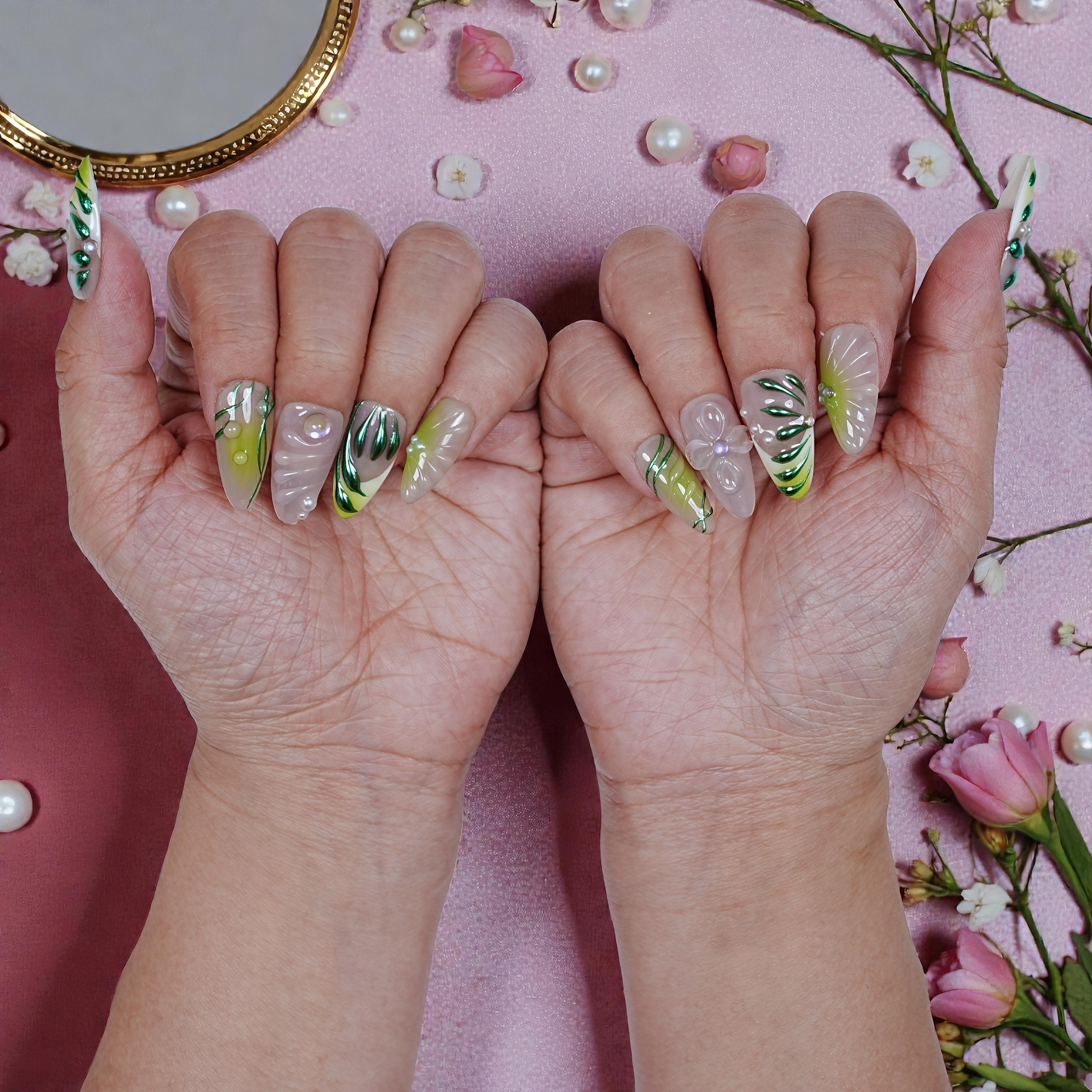Hands with long press on nails featuring green leaf and 3D flower designs on a pink background with pearls and pink flowers