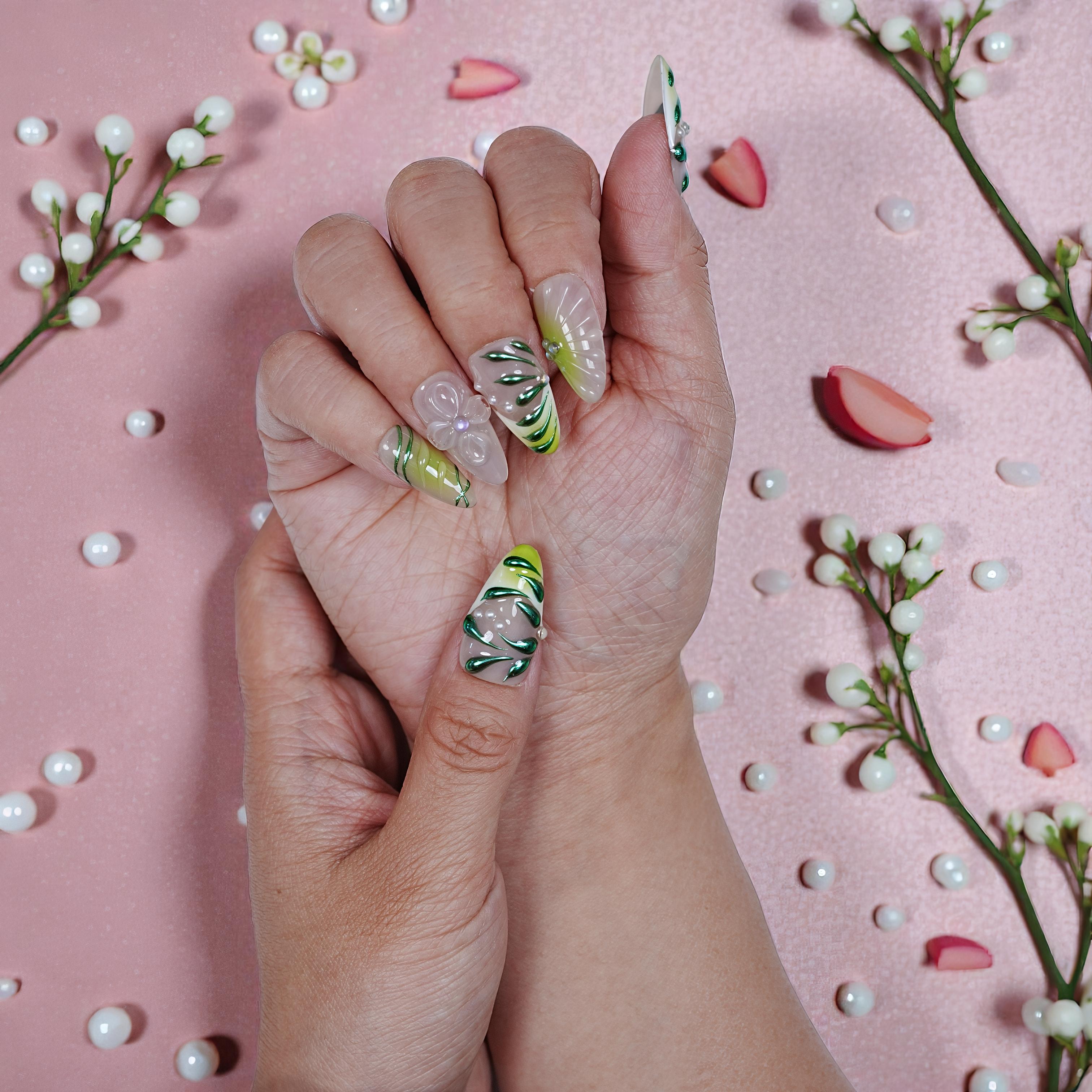 Hand with long, reusable press-on nails featuring green leaf designs and 3D floral accents over a pink background with decorative pearl-like beads and flower petals