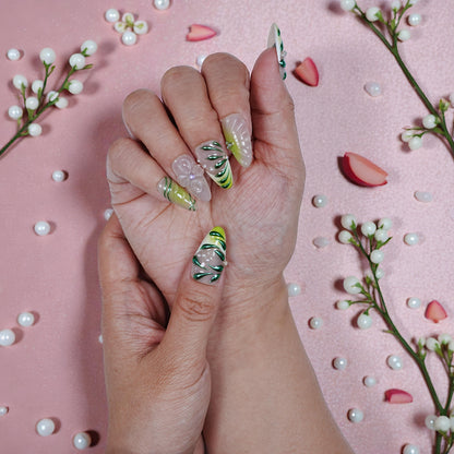 Hand with long, reusable press-on nails featuring green leaf designs and 3D floral accents over a pink background with decorative pearl-like beads and flower petals
