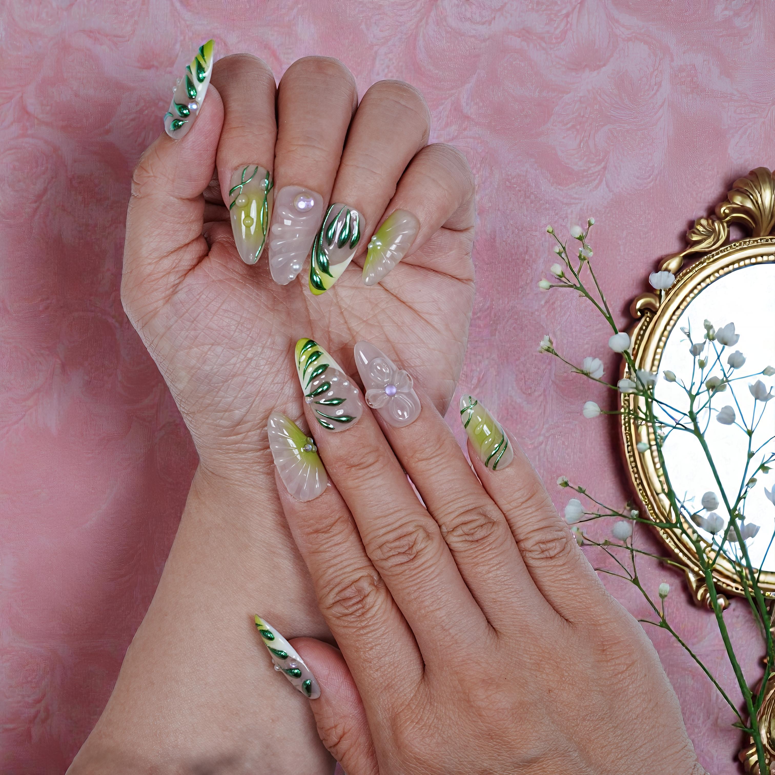 Hands showing long, handmade press on nails with green leaf designs, 3D floral accents, against pink textured background with gold mirror and white flowers