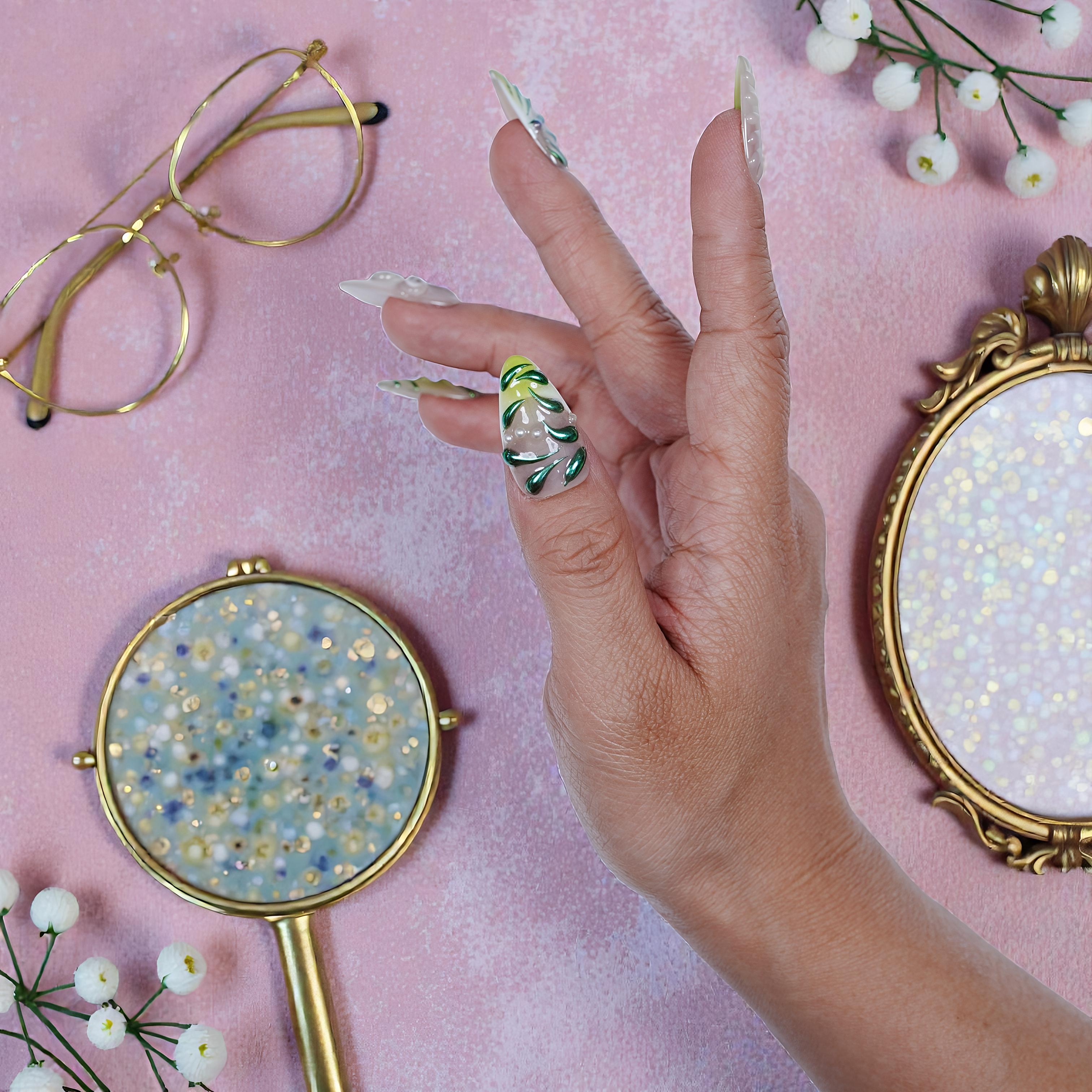 Hand with long handmade press on nails featuring green leaf designs on pink textured background with gold glasses and decorative mirrors
