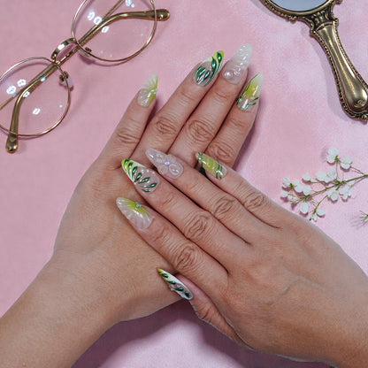 Hands with long green and white floral press on nails, styled with pearl and leaf designs on pink background