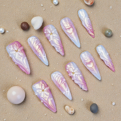 Long stiletto press on nails in pink and white chrome with 3D bows, hearts, and pearl accents displayed on sand with shells