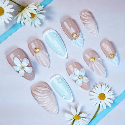 Flat lay of Blue Daisy Shell press on nails featuring sculpted white flowers, soft pink nude base, and baby blue tips with daisies and gold accents.