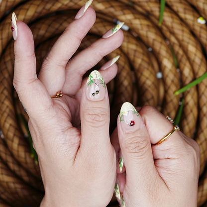 Thumb-focused hand pose with bumblebee art and grass linework over sheer nude polish.