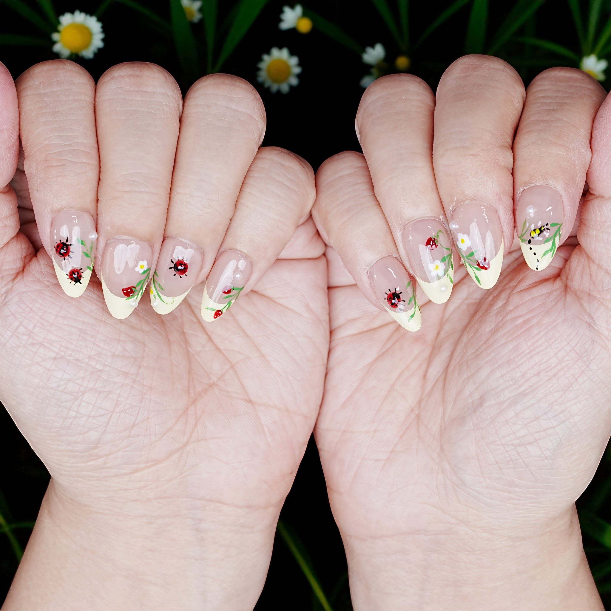 Both hands with ladybug press on nails in a spring setting, styled over daisy flowers and grassy background.